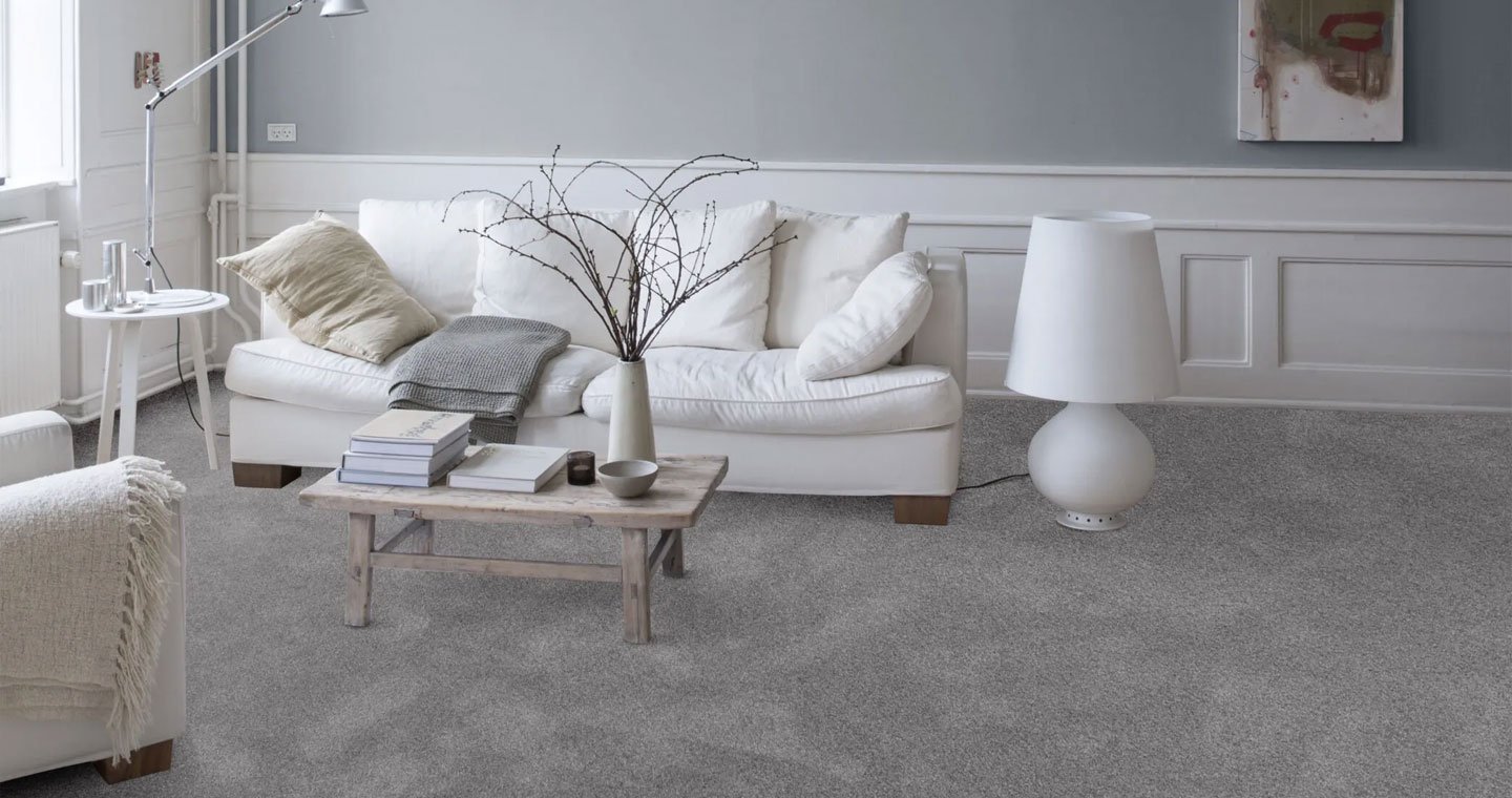 A living room with a white couch, a gray blanket, beige pillows, a wooden coffee table with books, a white diffuser, and a tall white lamp on a gray carpet, with white walls and a small window.