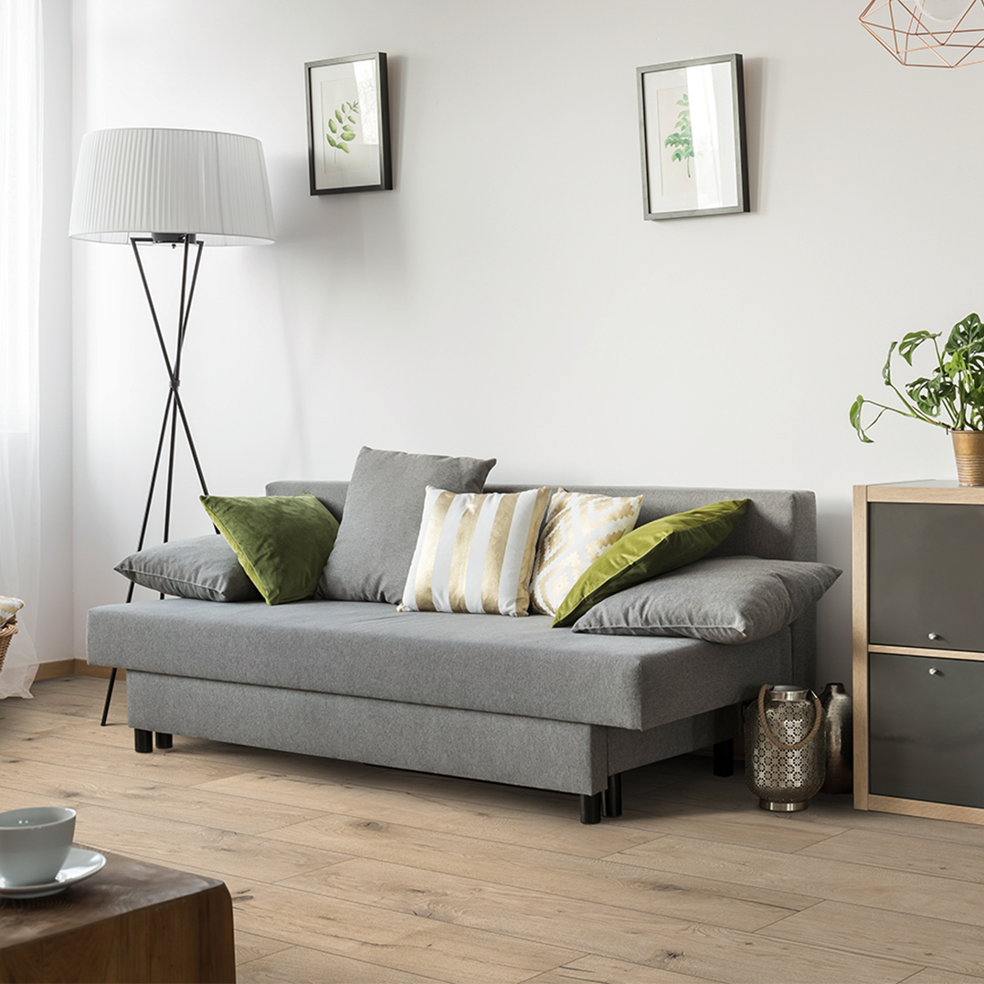 A modern living room with a gray sofa accented with green and patterned throw pillows, a tall white floor lamp, framed botanical prints on a white wall, a wooden and gray side table with a potted plant, and a wooden coffee table with a white cup and saucer.