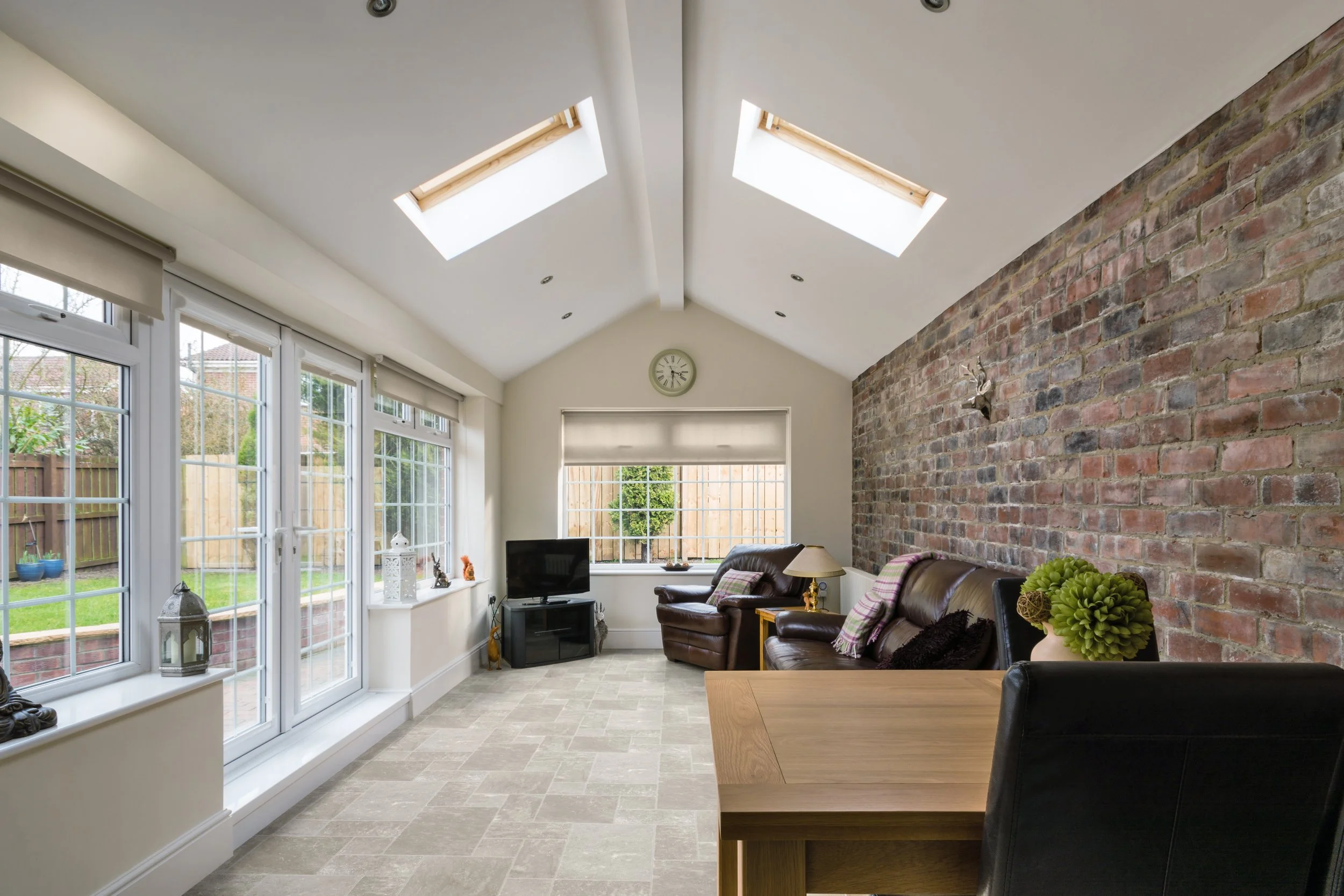 Living room with large windows, skylights, brick accent wall, leather sofa, armchair, TV, and decorative plants.
