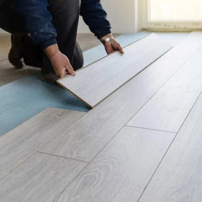 Person installing light-colored wooden flooring in a room, kneeling on the floor, handling a large plank.