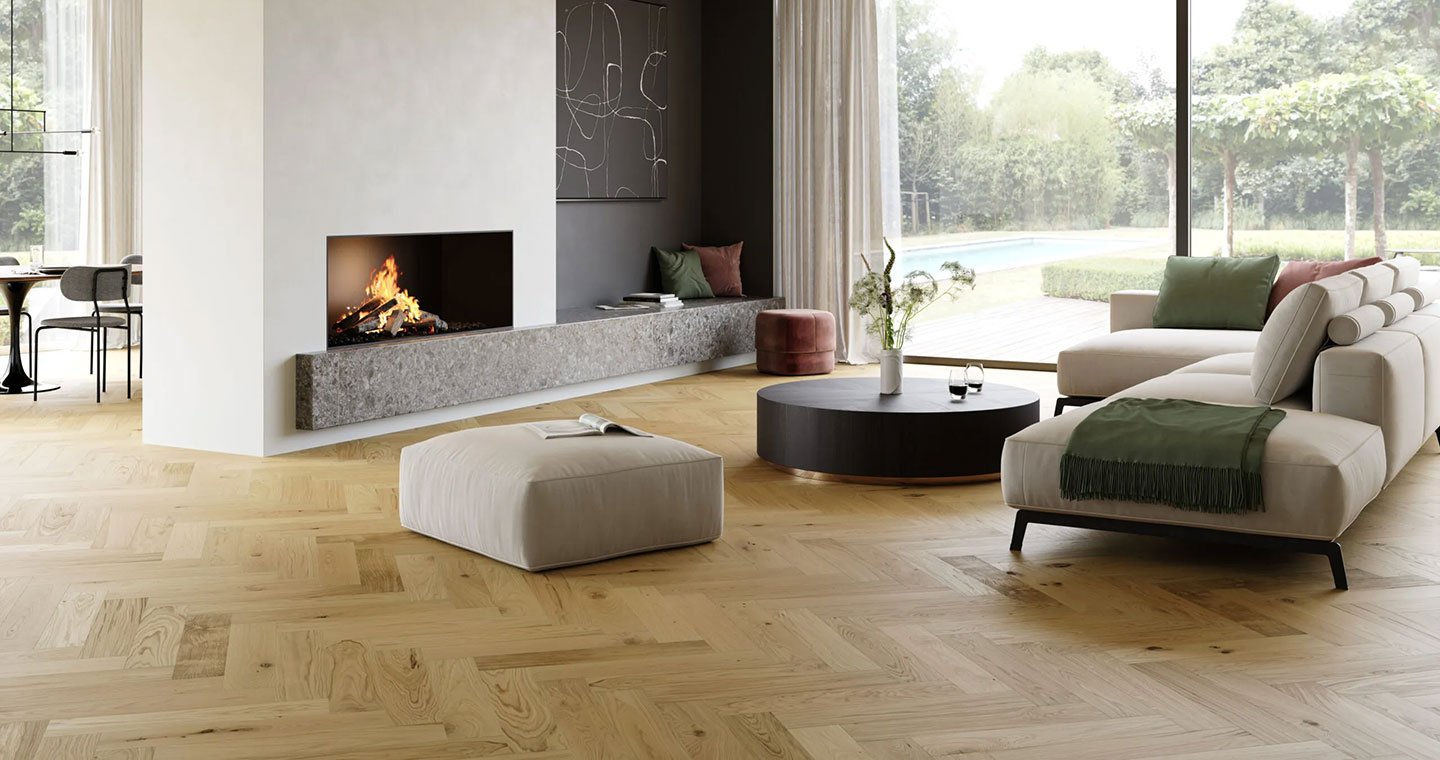 Modern living room with a fireplace, light wooden flooring, white sectional sofa with green and pink pillows, round black coffee table with a vase and glasses, large sliding glass door showing outdoor trees and grass, and a small ottoman with an open book.