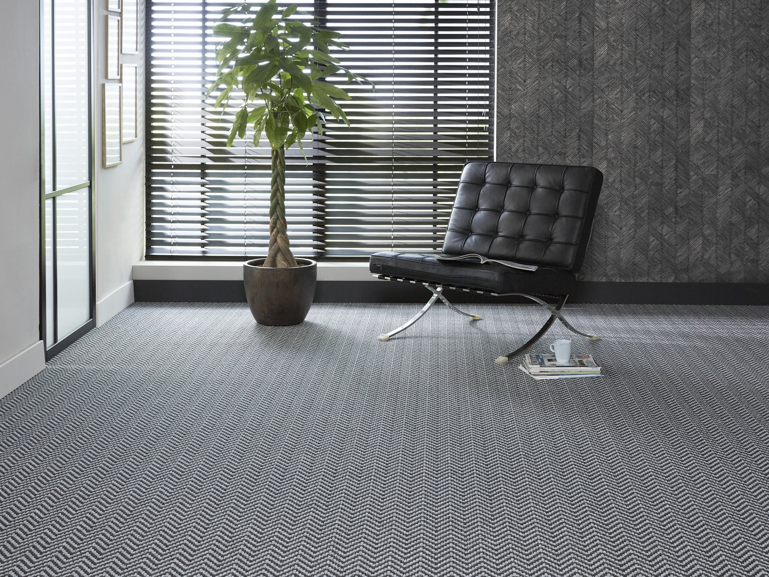 An office corner with a potted plant, a black lounge chair with a magazine, a mug, and newspapers on the floor, framed pictures on the wall, black window blinds, and textured gray carpet.