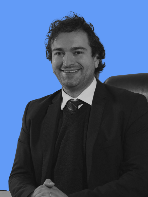 A man in a suit sitting in an office chair, smiling, with a blue background.