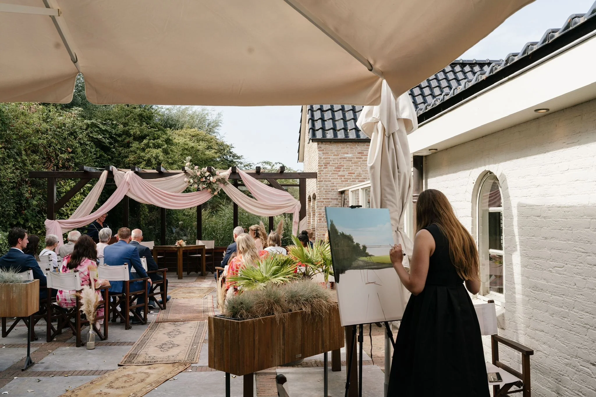 An outdoor wedding ceremony with guests seated under a decorated arch, and an artist painting a landscape on an easel.