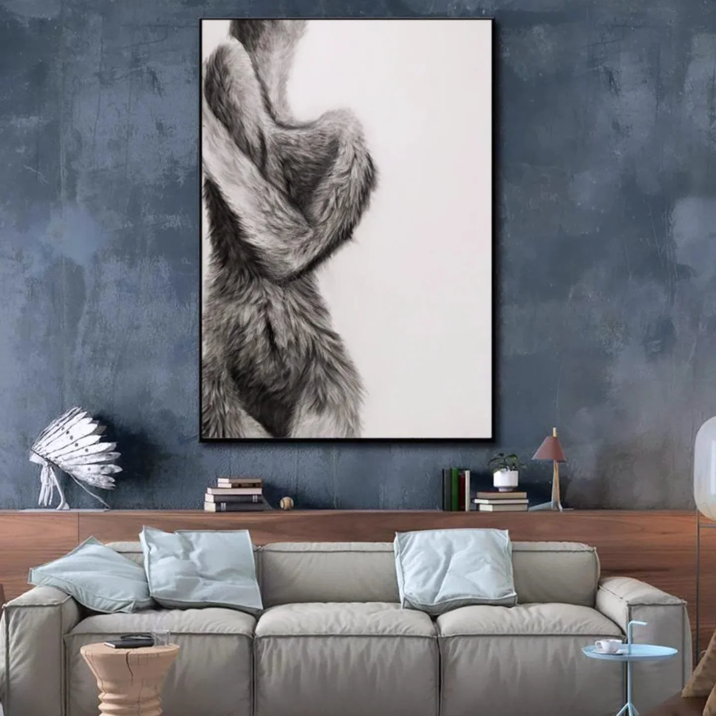 A living room with a beige sofa, pillows, a wooden side table, and a large framed black and white photograph of a furry animal, likely a wolf or dog, hanging on a textured dark blue wall. There are decorative items like a small white sculpture, a stack of books, a potted plant, and a table lamp on the shelf behind the sofa.