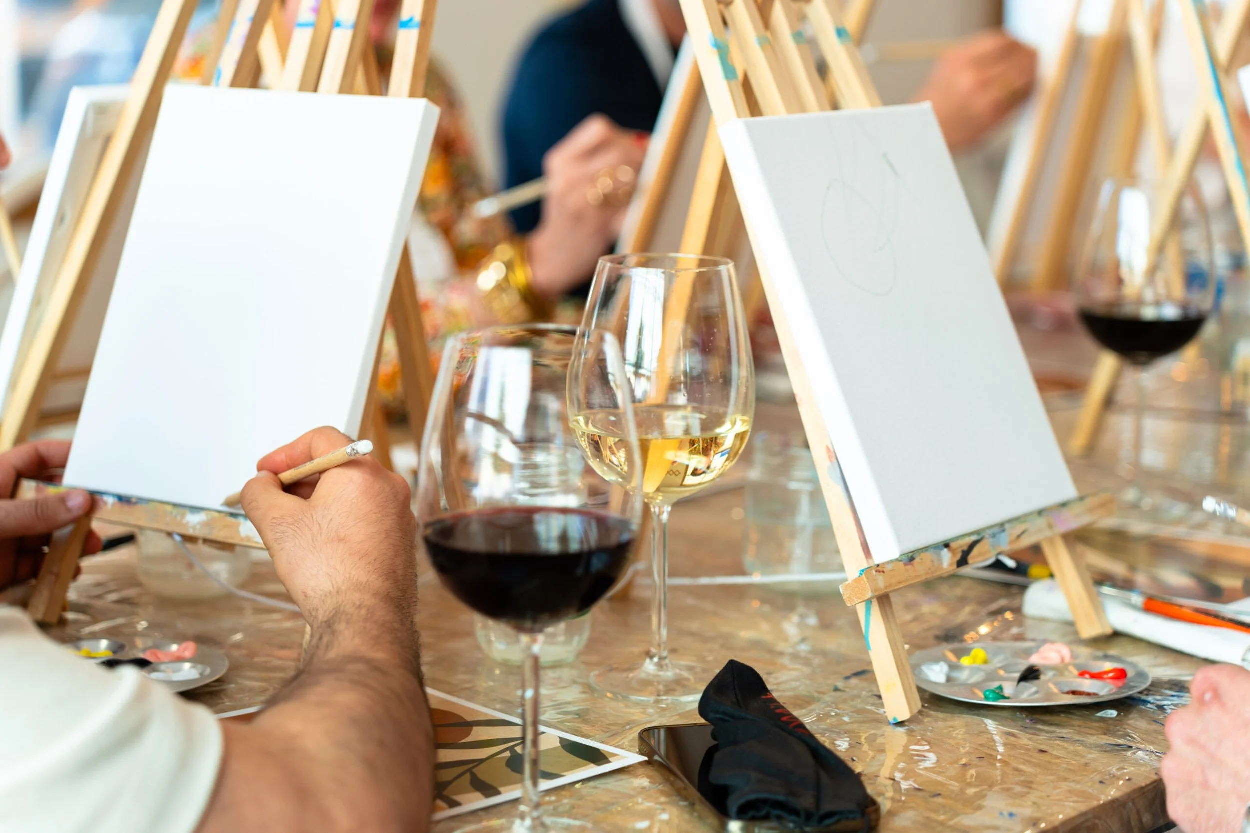 People participating in a group art activity, painting on blank canvases on small easels at a table with glasses of red and white wine.