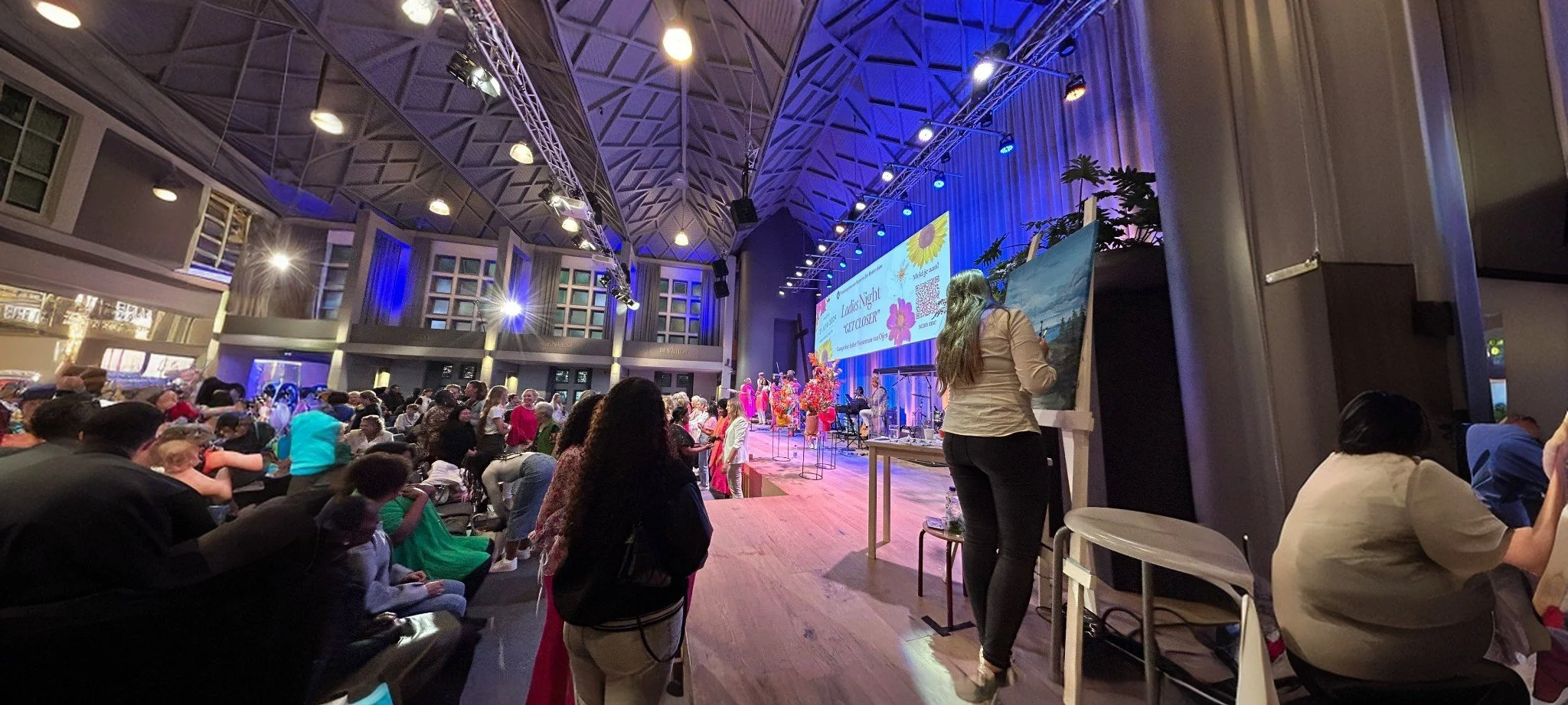 A large indoor event with many people gathered, watching a performance on stage. The stage is decorated with bright colors, flowers, and a large screen displaying event information. The venue features a high, arched ceiling with lighting and rigging.