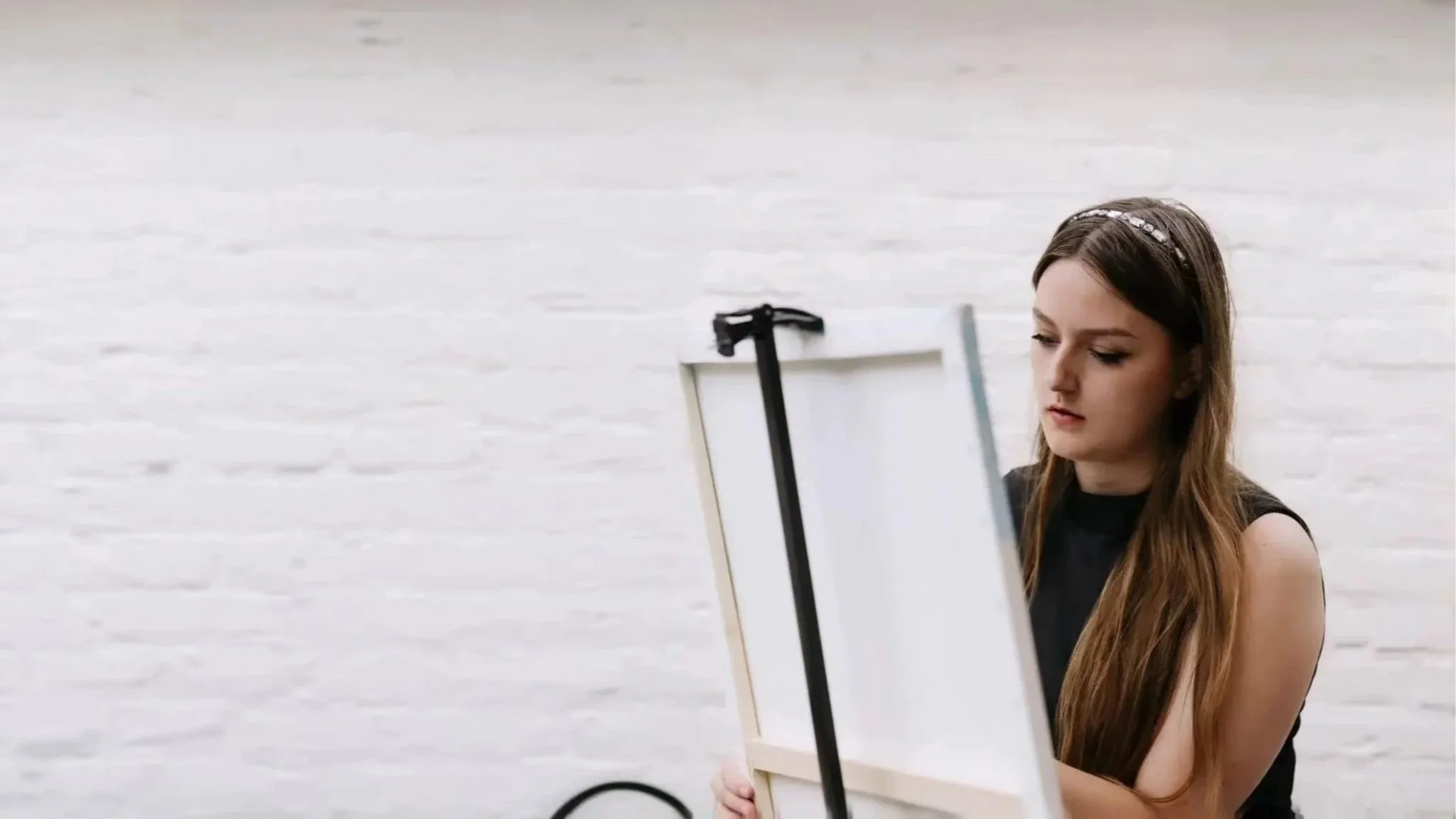 Young woman with long brown hair and a headband painting on a blank canvas set on an easel against a white brick wall.