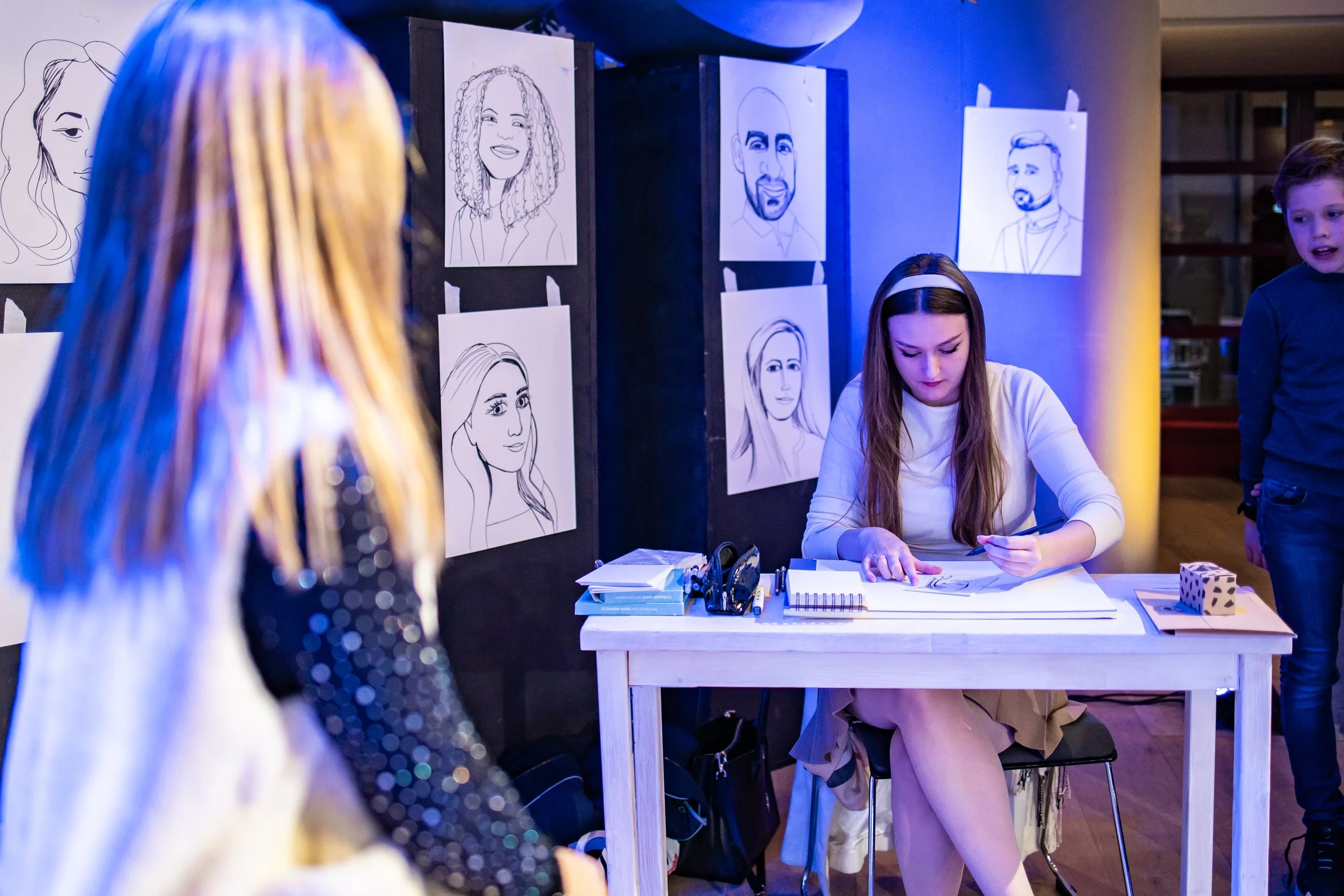 Artist drawing caricature portraits at an art event in a room decorated with line art drawings on the wall.