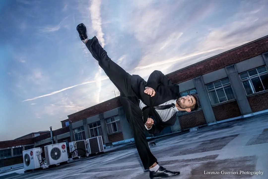 A man in a business suit performing a high kick on a rooftop under a cloudy sky.