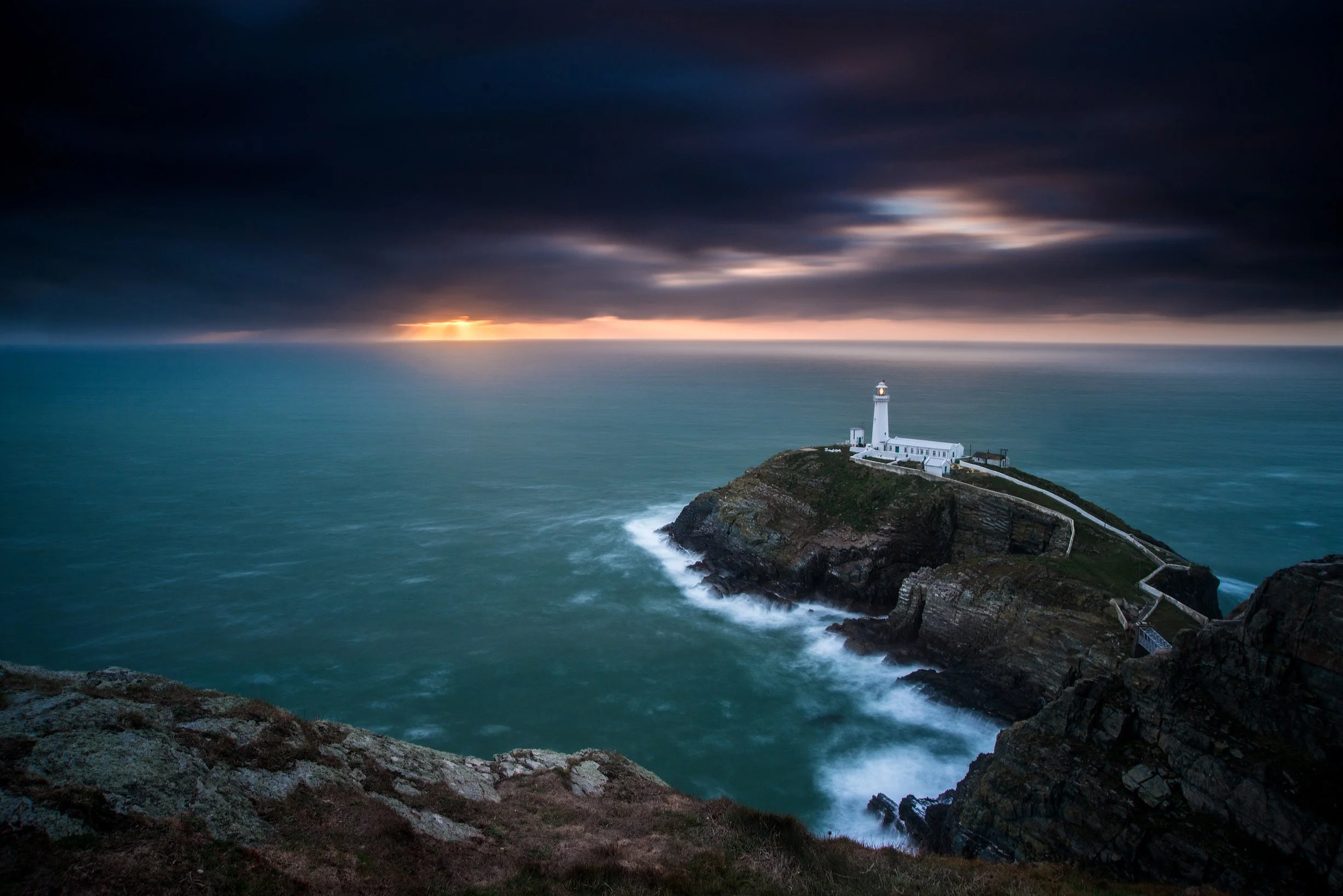 A lighthouse on a rocky island at sunset with dark clouds and choppy ocean waves.