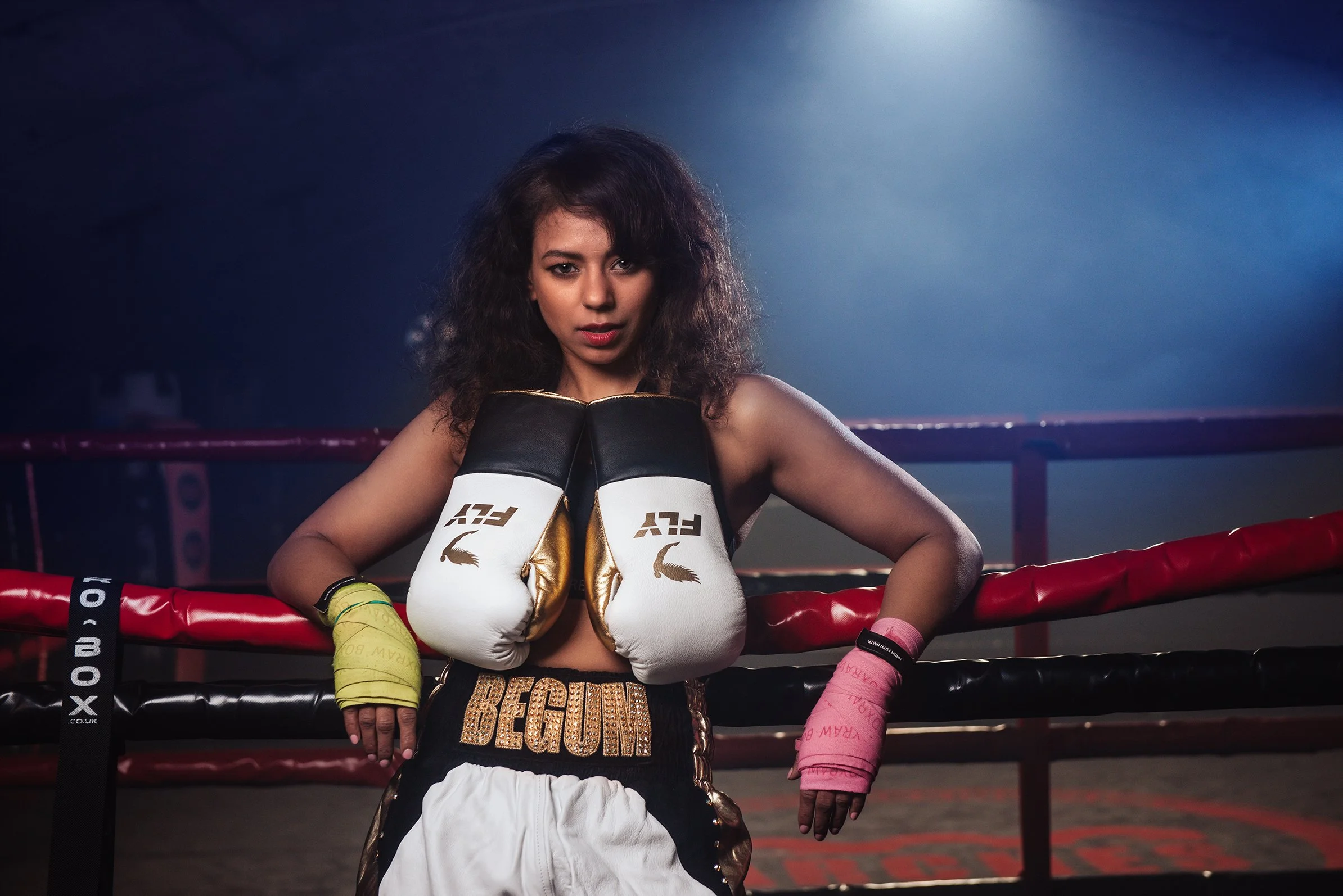 Female boxer with curly hair leaning on boxing ring ropes, wearing boxing gloves and boxing trunks with "BEGIN" written on them, in a dimly lit boxing gym.