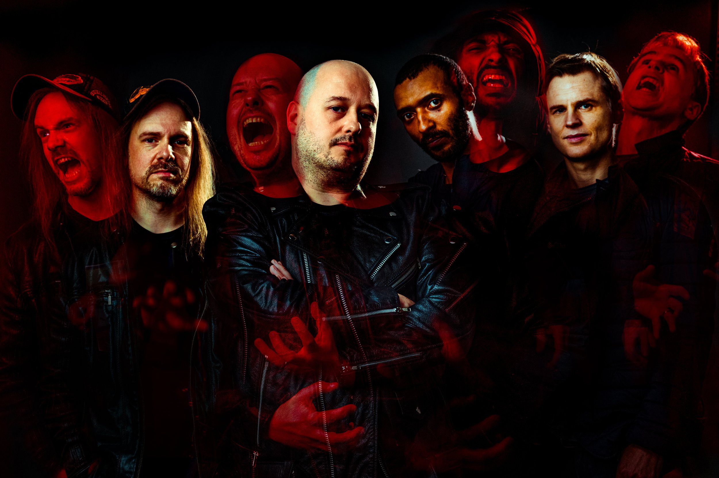 Group of eight men in dark clothing, some with arms crossed, some with angry or intense facial expressions, in a dark, red-lit setting.