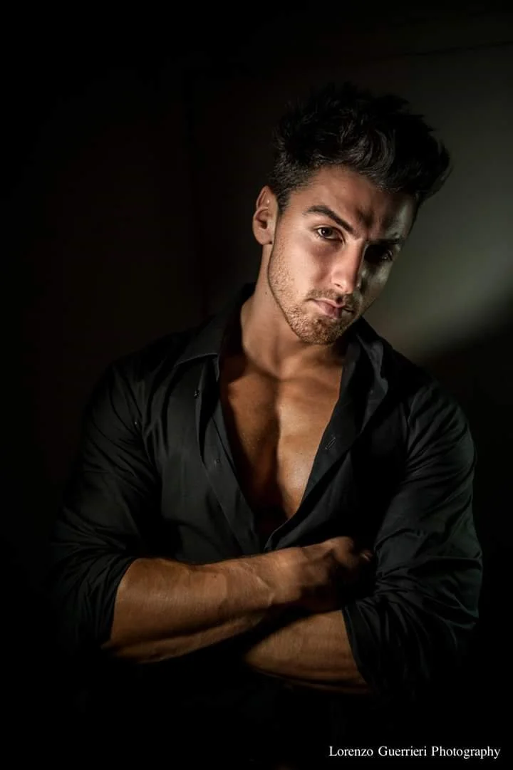 A young man with dark hair and stubble, wearing a black shirt with the top few buttons open, standing against a dark background with a light source illuminating his face and partly his chest.