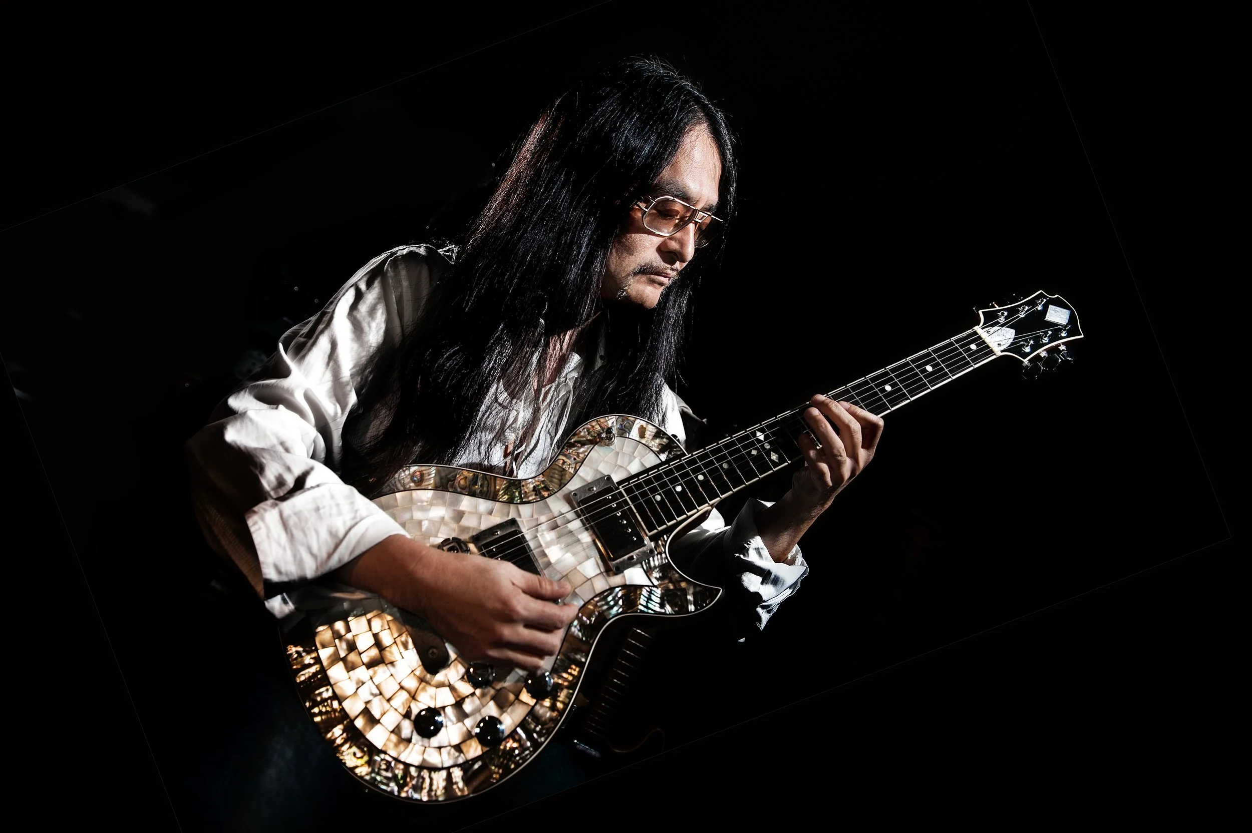 A man with long black hair, wearing glasses and a white shirt, playing an intricately designed electric guitar in a dark setting.