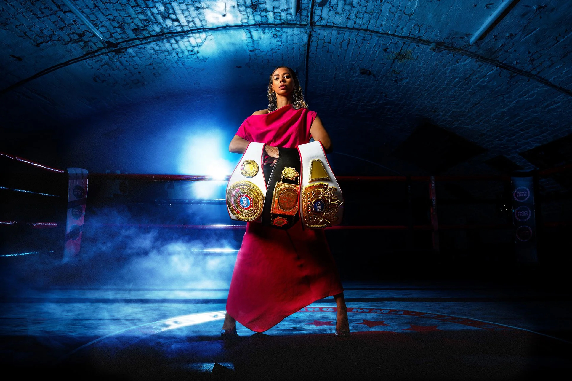 A woman standing in a boxing ring, holding multiple championship belts, with a spotlight behind her in a dimly lit gym.