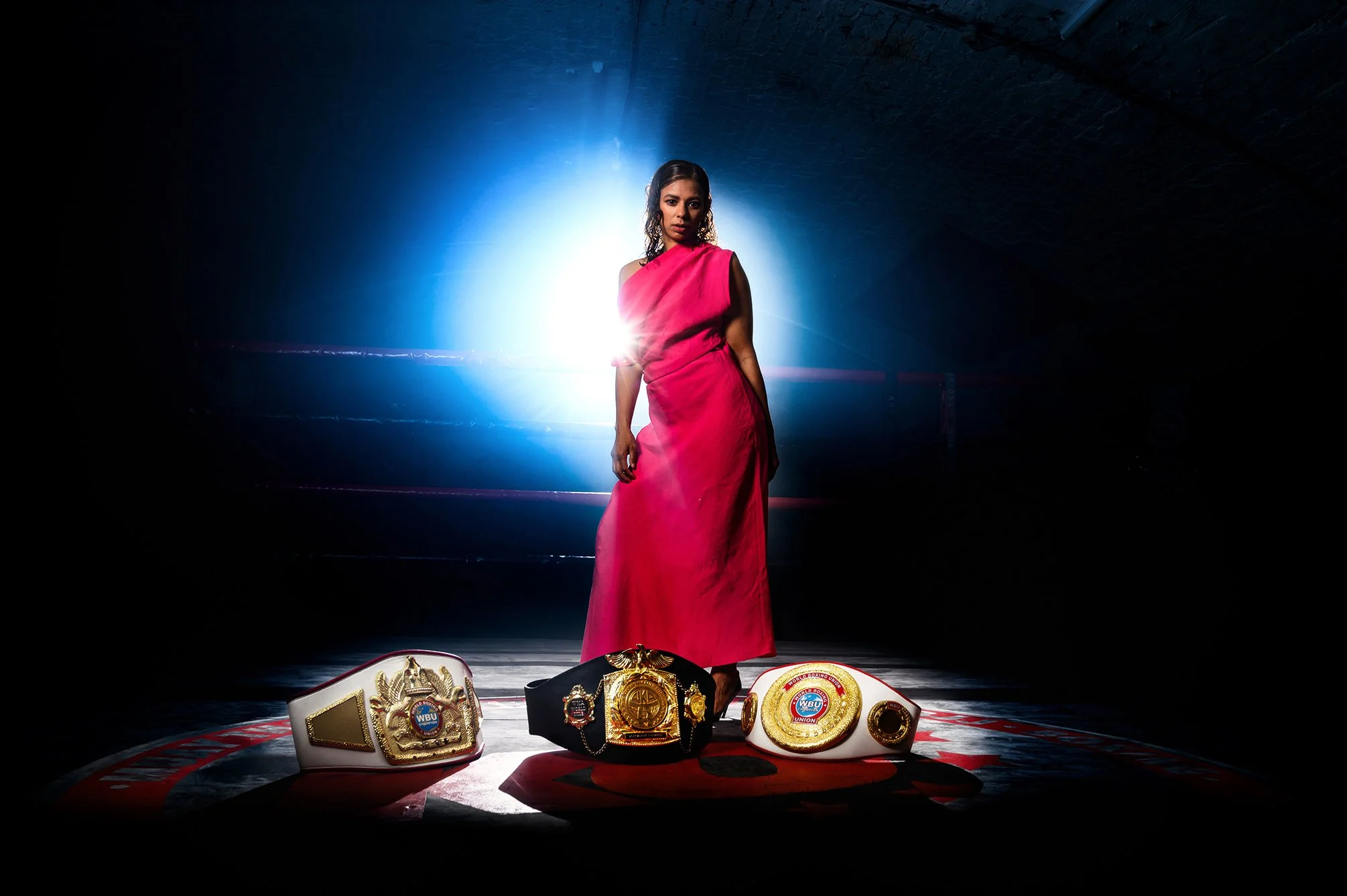 A woman in a pink dress standing on a boxing ring with four championship belts on the floor in front of her, illuminated by a bright light behind her.