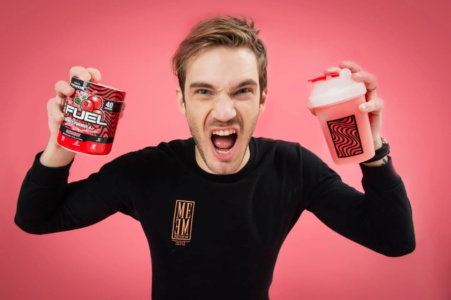 Young man with short hair and a black shirt holding a container of pre-workout supplement in his left hand and a shaker bottle in his right hand, screaming with excitement against a pink background.
