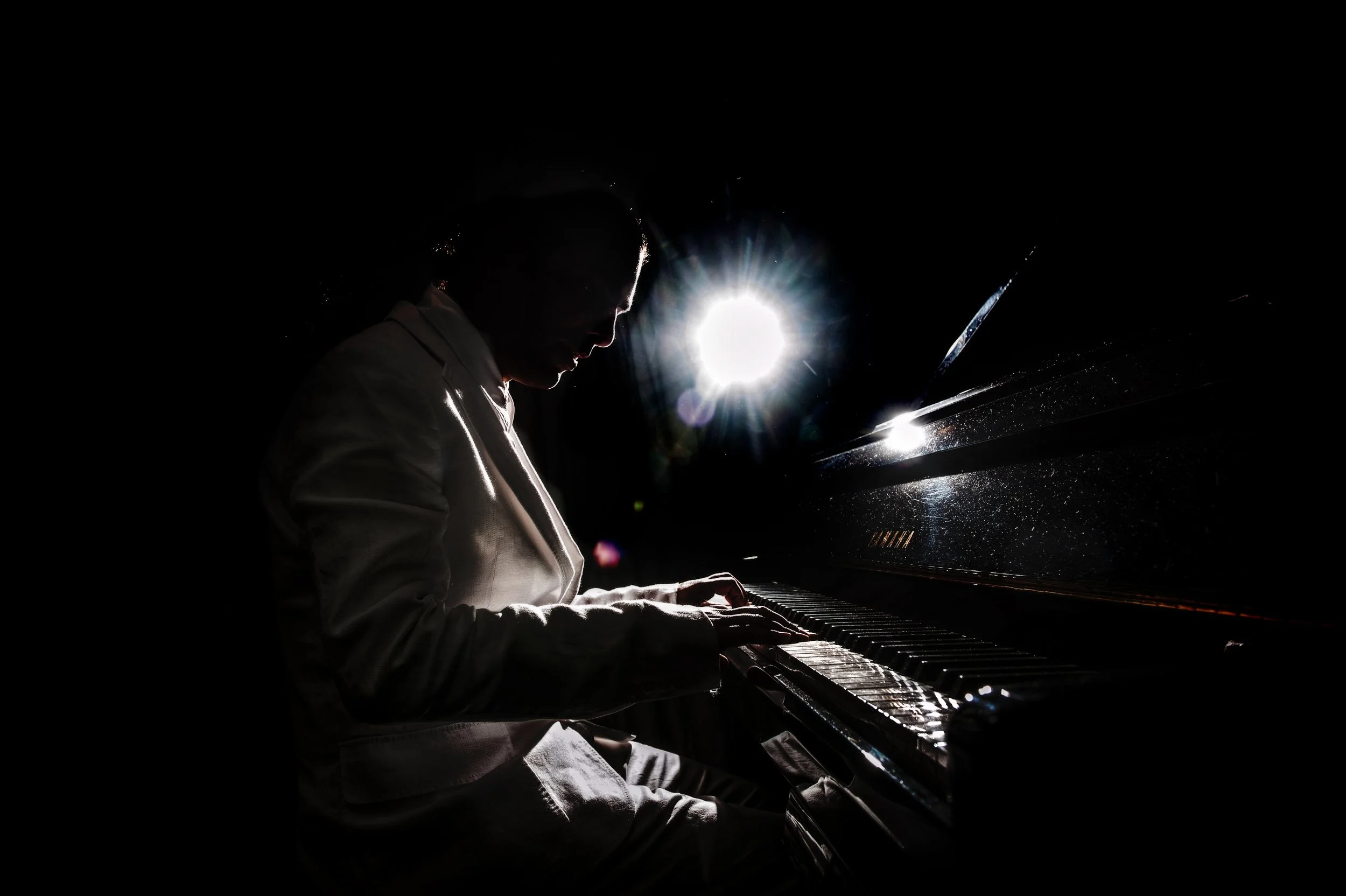 A person playing a grand piano in a dark setting with sunlight coming from behind.