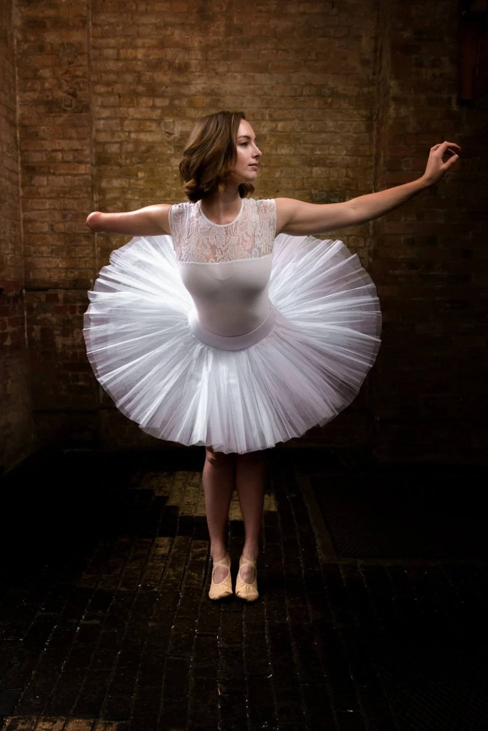A ballerina standing on a dark wooden floor with brick walls in the background. She is wearing a white leotard with lace on the upper part and a tutu, with her arms extended outward in a ballet pose.