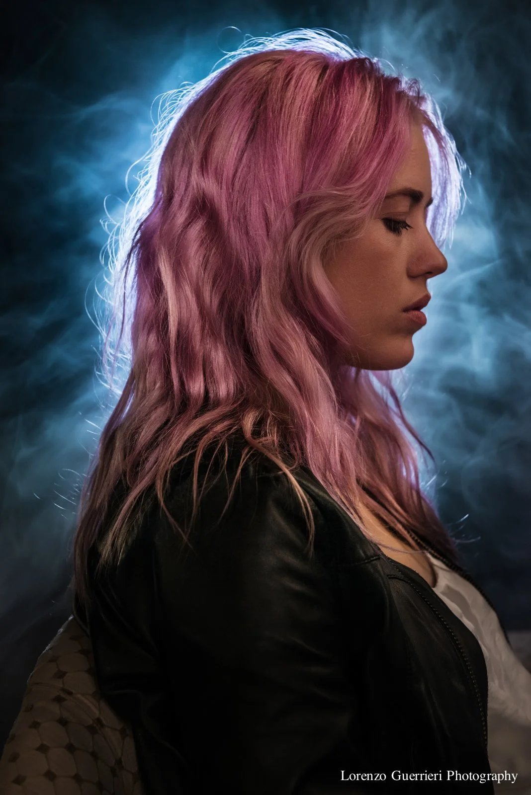 Side profile of a woman with pink hair, closed eyes, and wearing a black leather jacket against a smoky blue background.