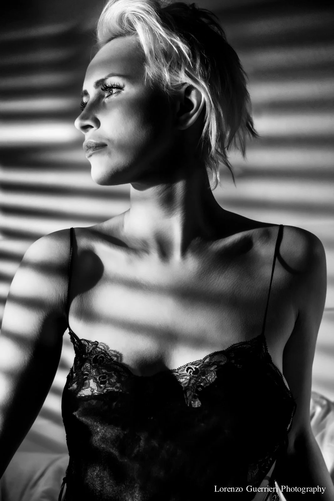 Black and white photo of a woman with short, light-colored hair, wearing a lace camisole, gazing to the side with patterned shadows cast across her face and shoulders.