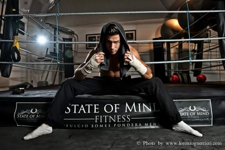 Male boxer on a boxing ring with a serious expression, sitting in a wide stance, wearing a hoodie and hand wraps, with gym equipment in the background.