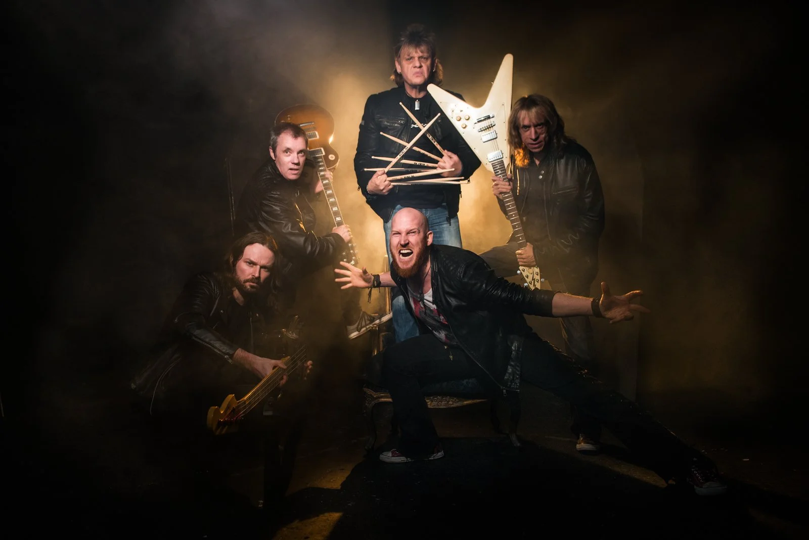 Group of six male rock musicians posing dramatically with guitars and drumsticks in a smoky, dark setting, illuminated by yellowish lighting.