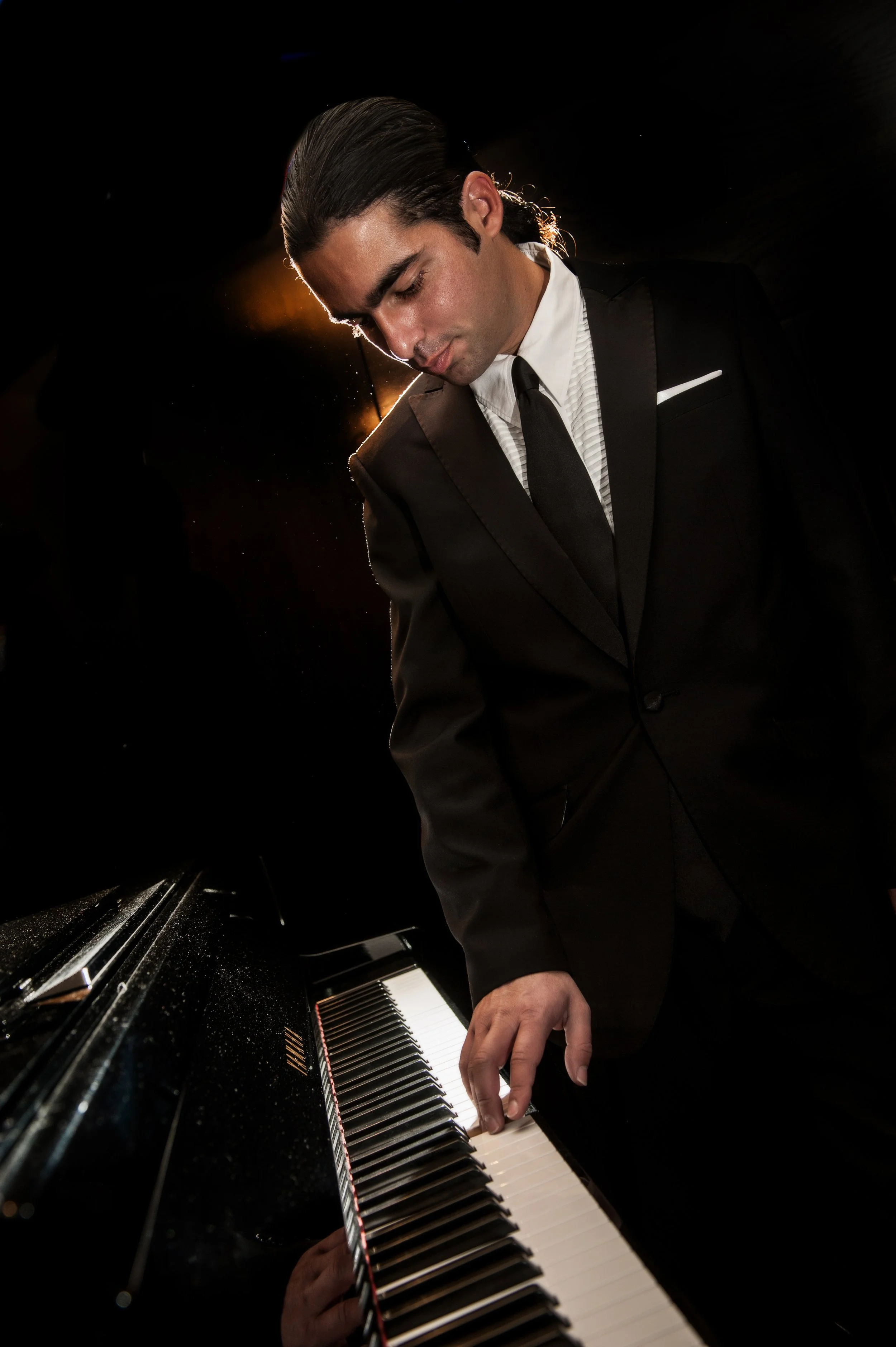 A man in a black suit and tie playing a grand piano, with dramatic lighting highlighting his face and hair.