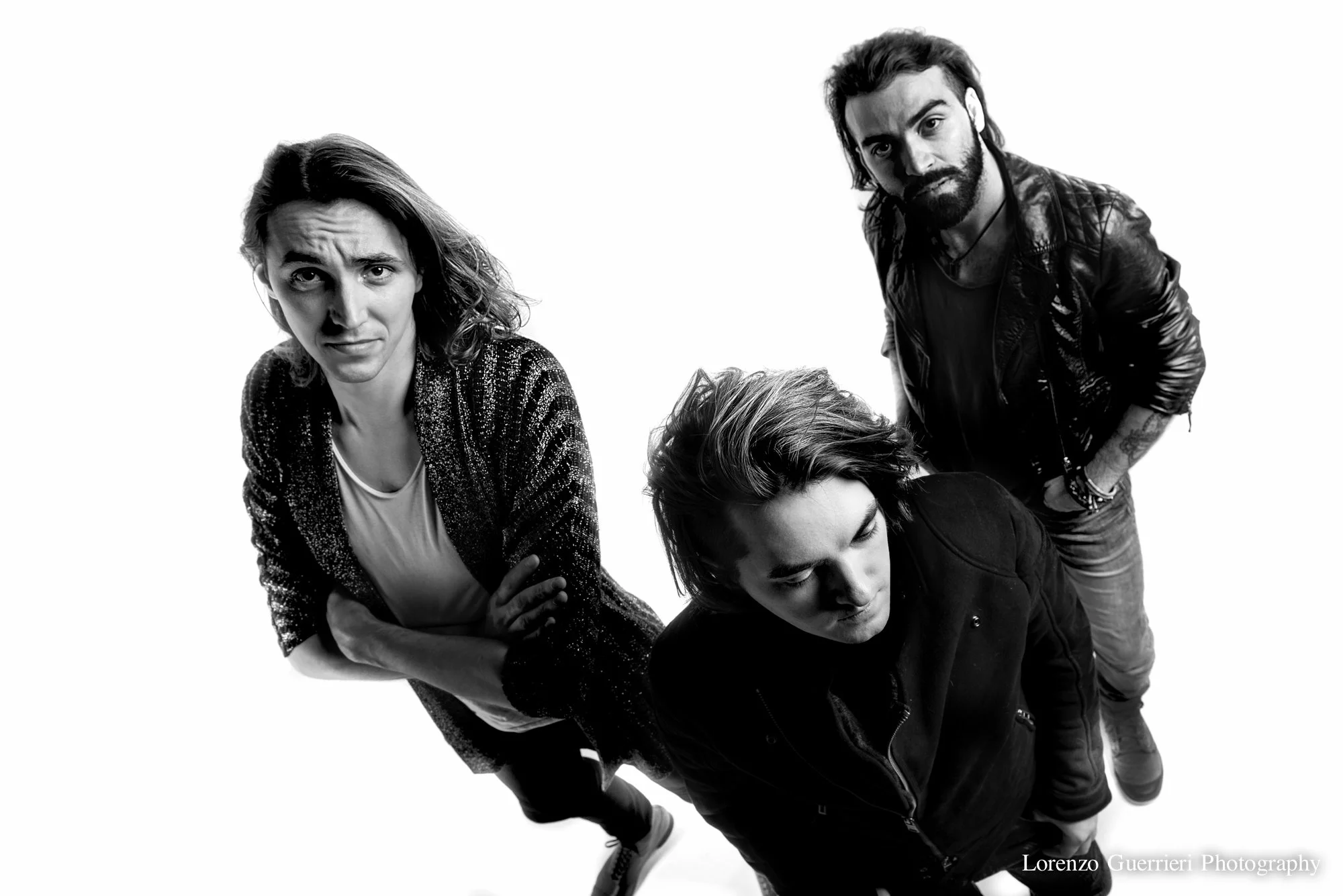 Black and white photo of four people, three men and one woman, looking up at the camera. One woman with wavy hair is on the left, making a confused or concerned face. The man on the right has a beard and is wearing a leather jacket. The man in the ce