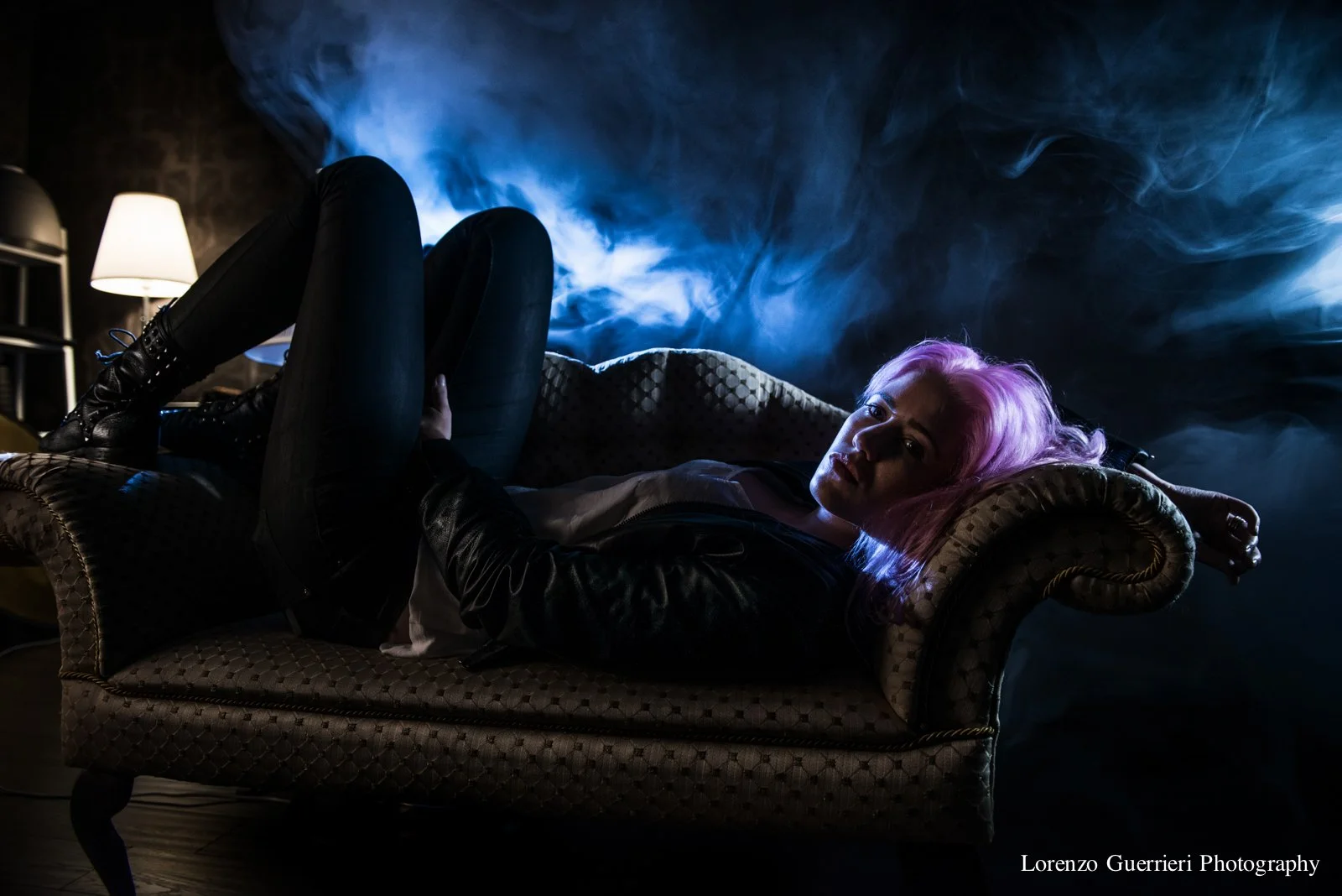 A woman with pink hair lies on a vintage style upholstered chaise lounge in a dark room with blue smoky atmosphere, illuminated by a nearby lamp.