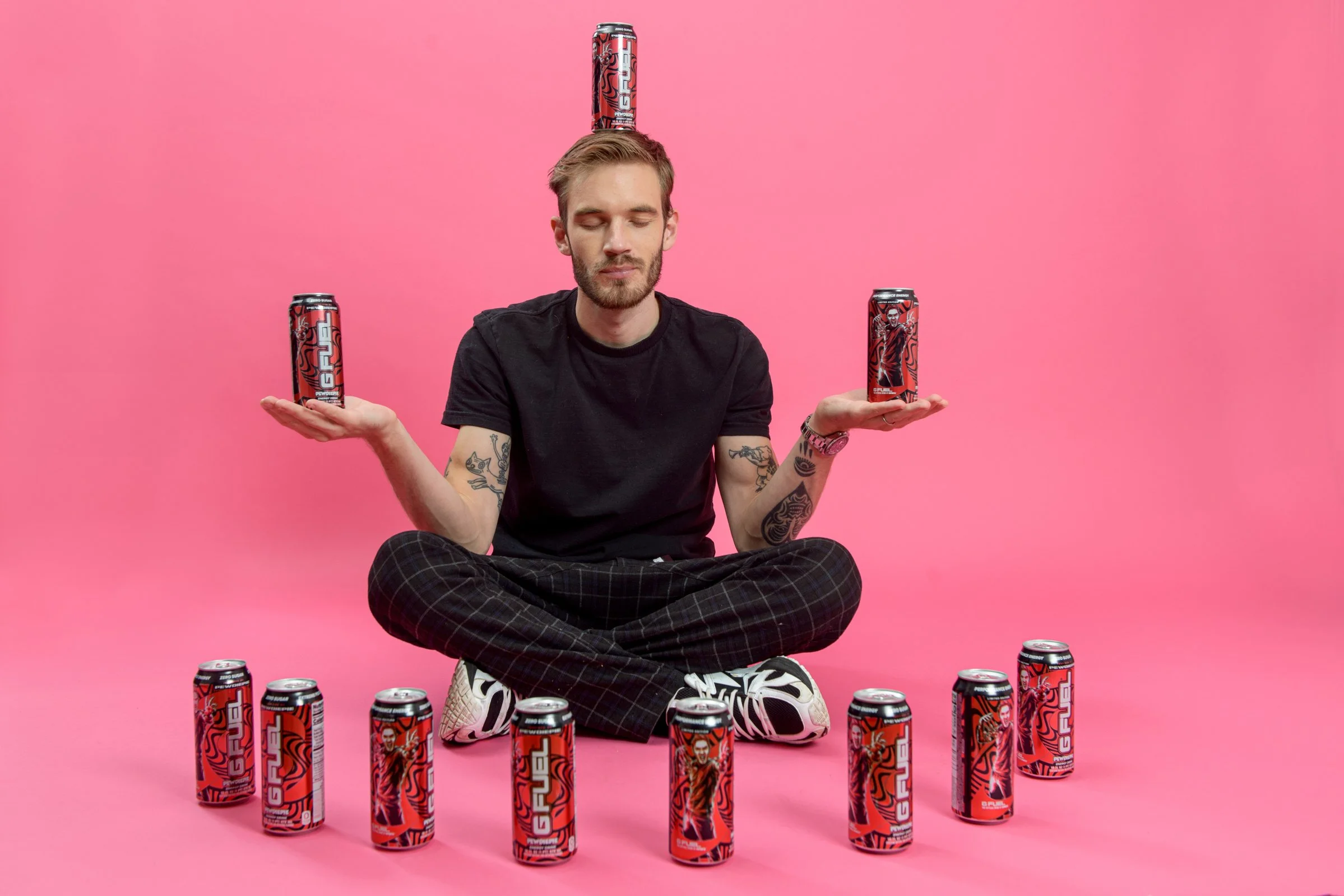 A man with tattoos sitting cross-legged on a pink background, balancing six cans of Red Bull energy drink with three on the ground and three balanced on his hands and head.