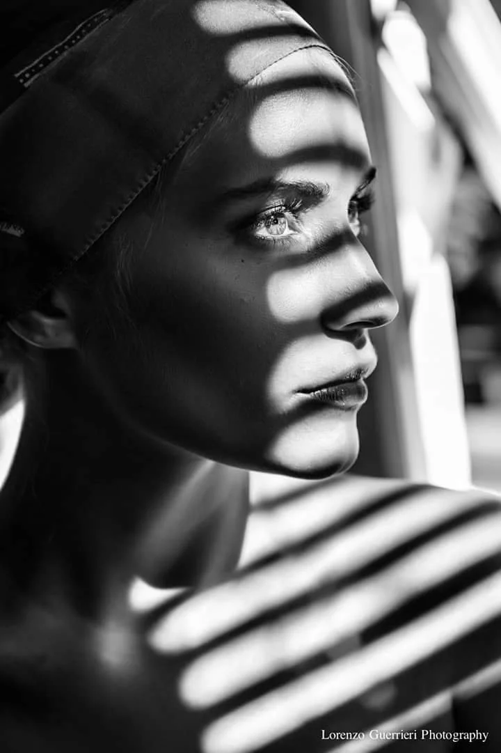 A black and white close-up of a woman looking out a window, with sunlight creating striped shadows across her face and shoulder, wearing a cap and a striped shirt.