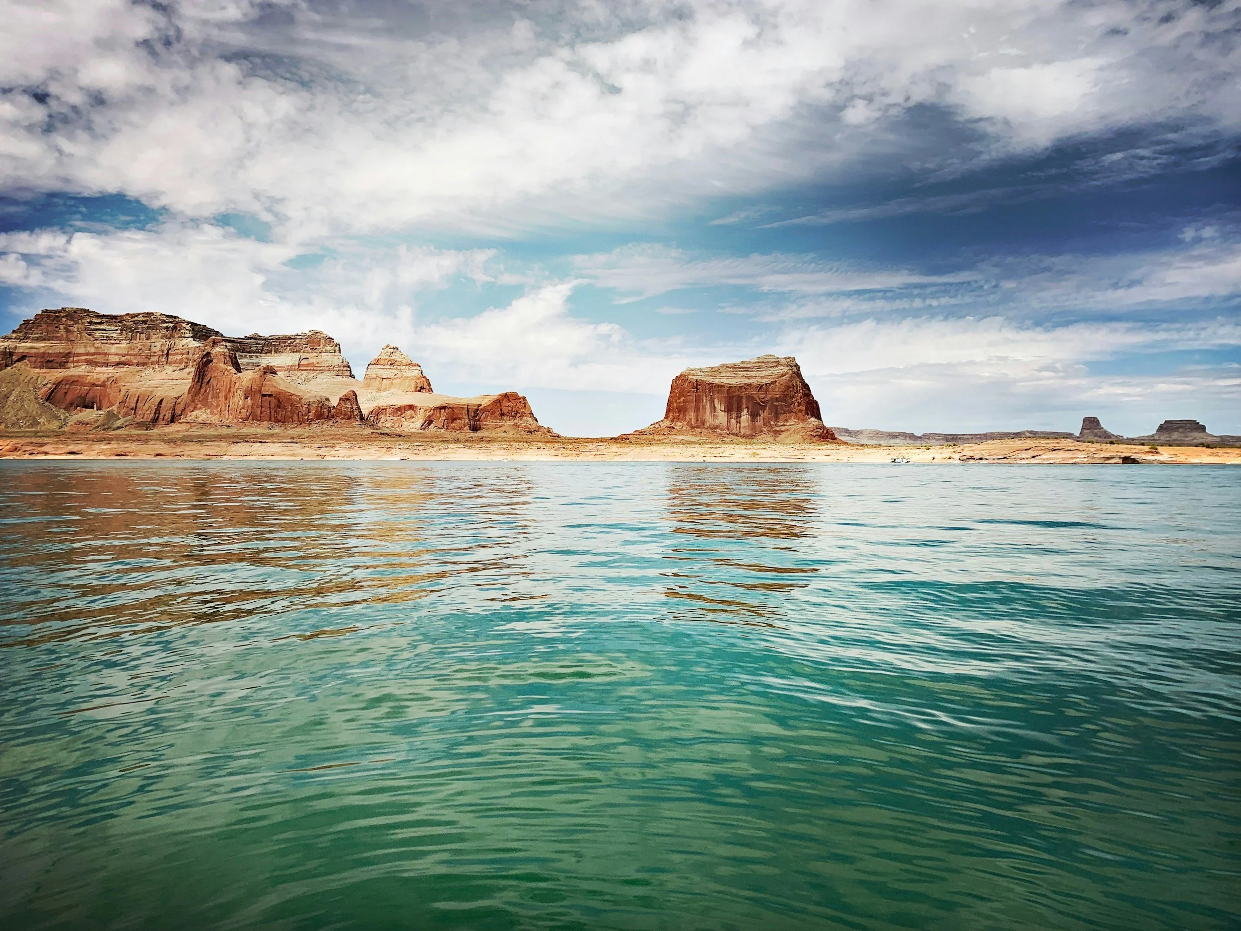 Scenic view of a large body of water with red rock formations and cliffs in the distance under a partly cloudy sky.