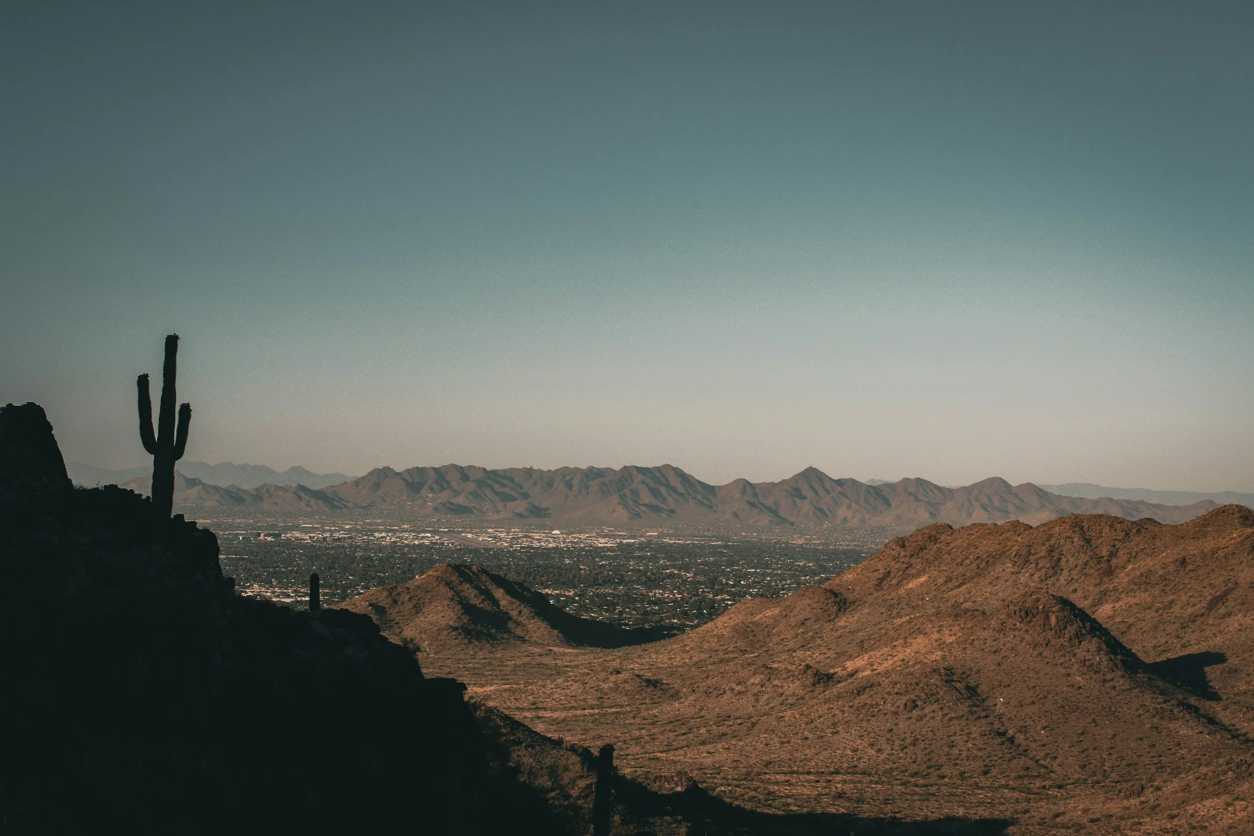 Desert mountain landscape with a saguaro cactus on the left foreground, brown rolling hills, and a distant mountain range under a clear sky.