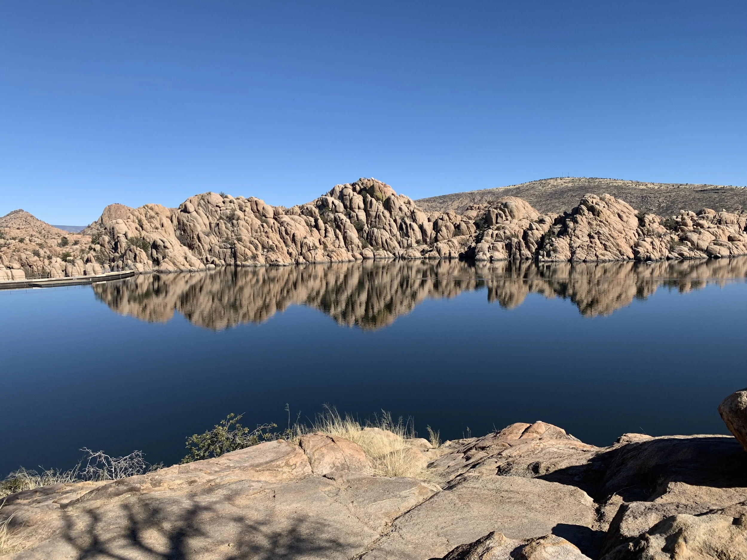 A rocky lakeshore with calm water reflecting rugged, beige-colored mountains under a clear blue sky.