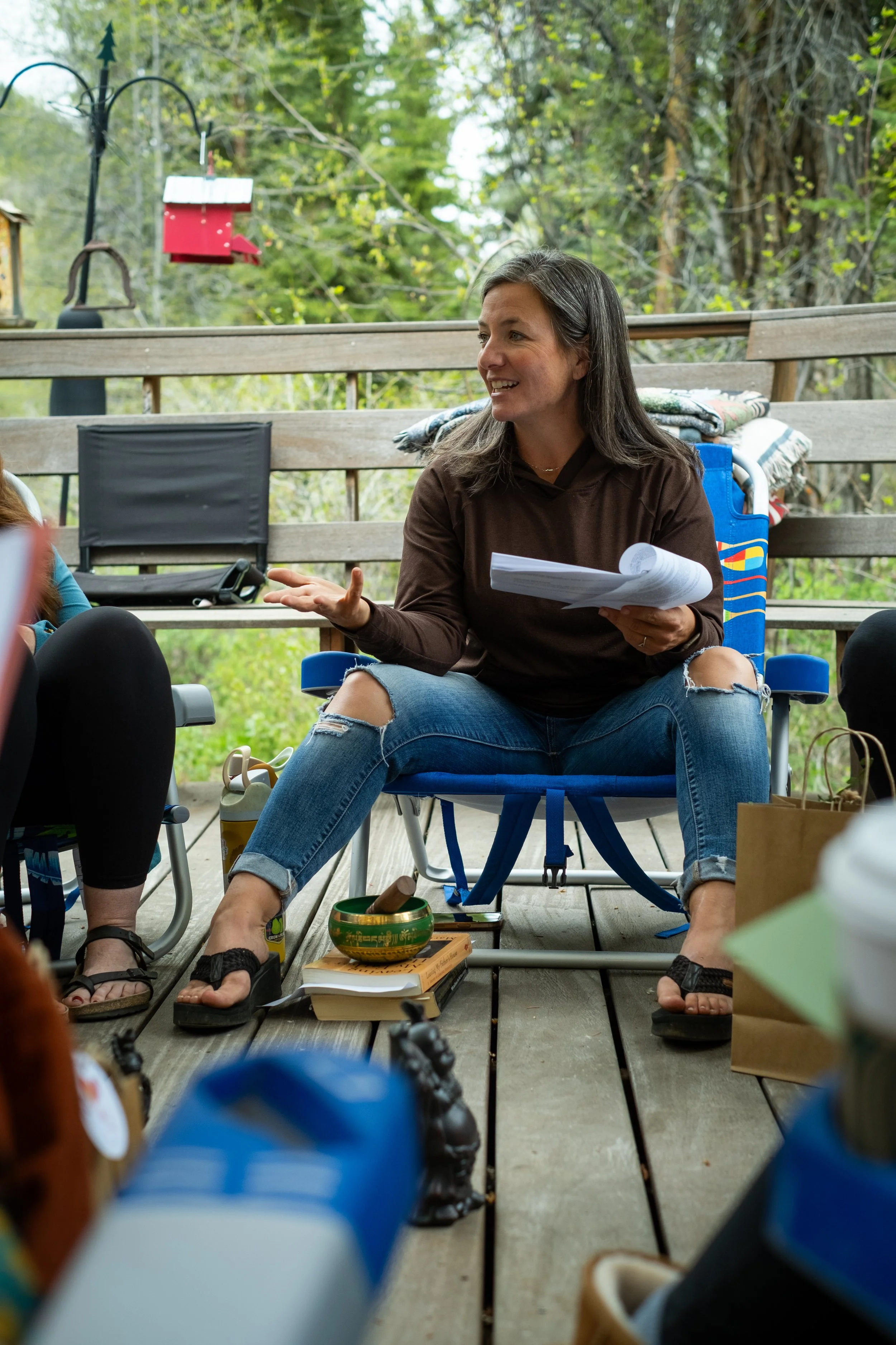 A woman with long dark hair, wearing a brown hoodie and ripped jeans, is sitting on a chair on a wooden porch, holding papers and talking. There are other chairs, books, a singing bowl, and a yellow thermos around her. The porch has a background of green trees and bird feeders.