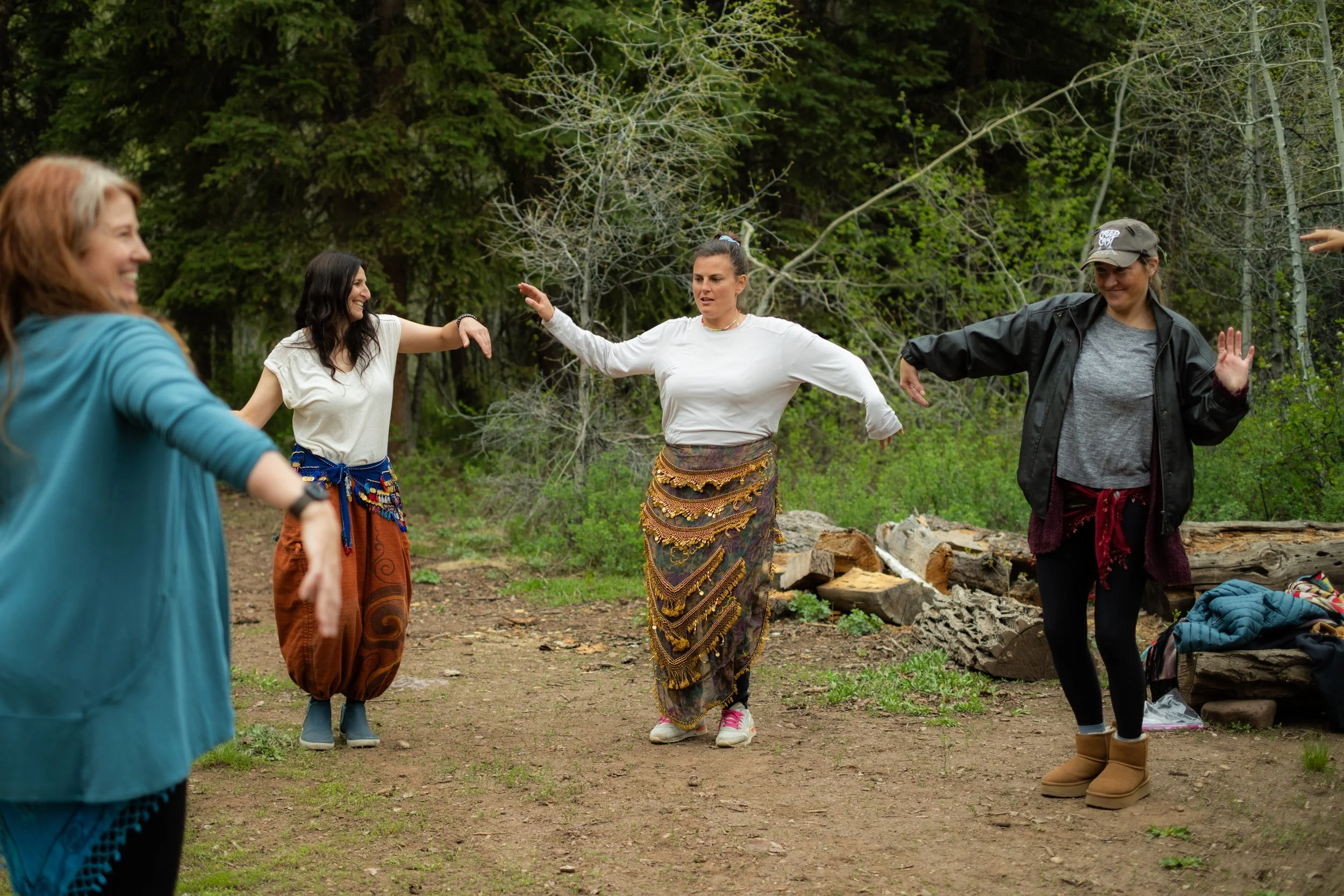 Four women dancing outdoors in a wooded area, dressed in casual and bohemian style clothing.