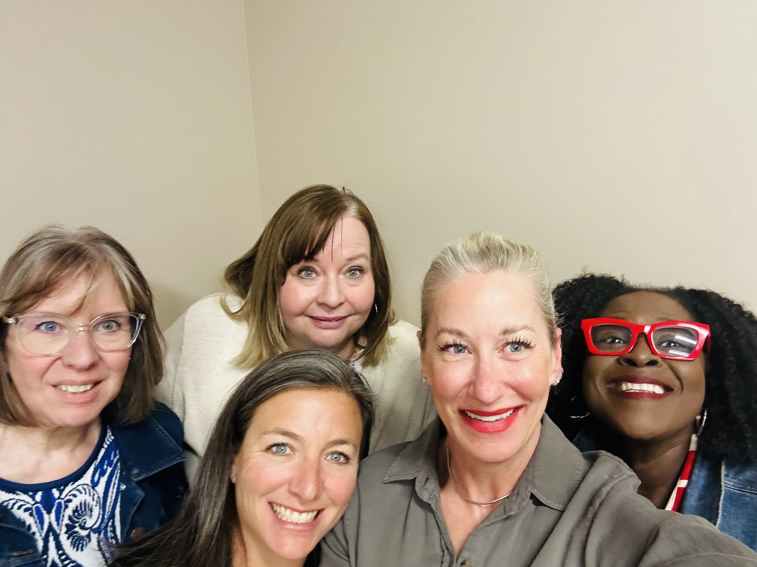 Group of women smiling and posing for a photo indoors against a plain wall.