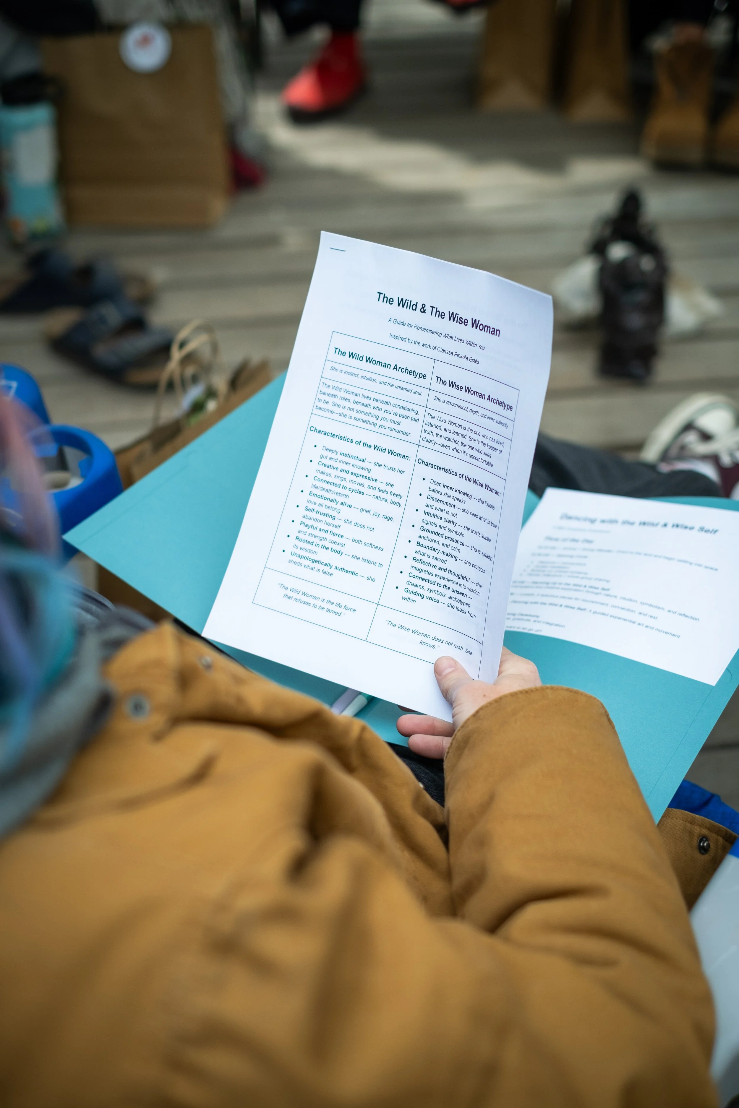 Person wearing a brown jacket holding a printed pamphlet that compares 'The Wild & The Wise Woman' archetypes, with shoes and other items on a wooden table in a setting that looks like a marketplace or workshop.