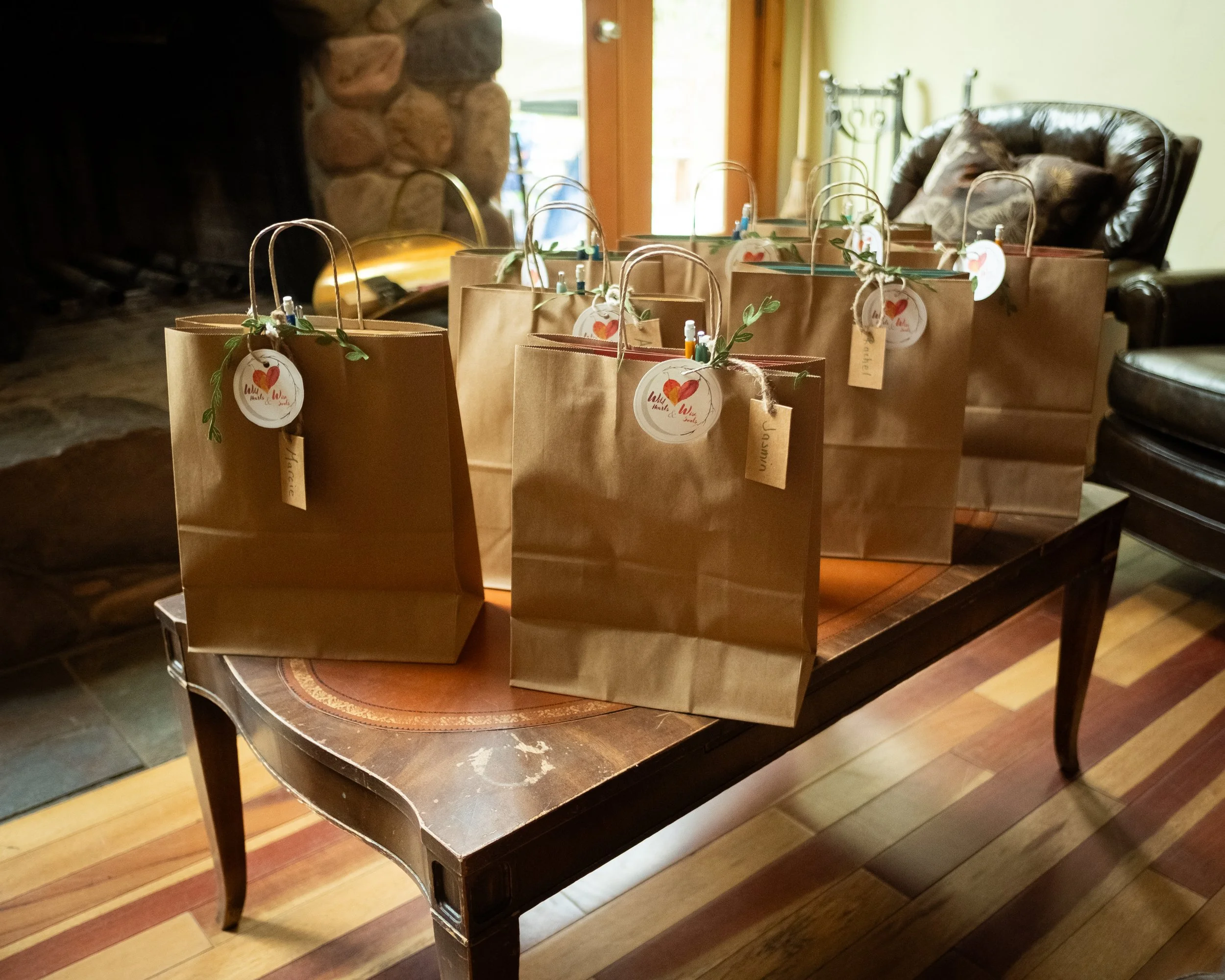 Brown paper gift bags with handles and tags, decorated with greenery, lined up on a wooden table in front of a fireplace and a leather armchair.