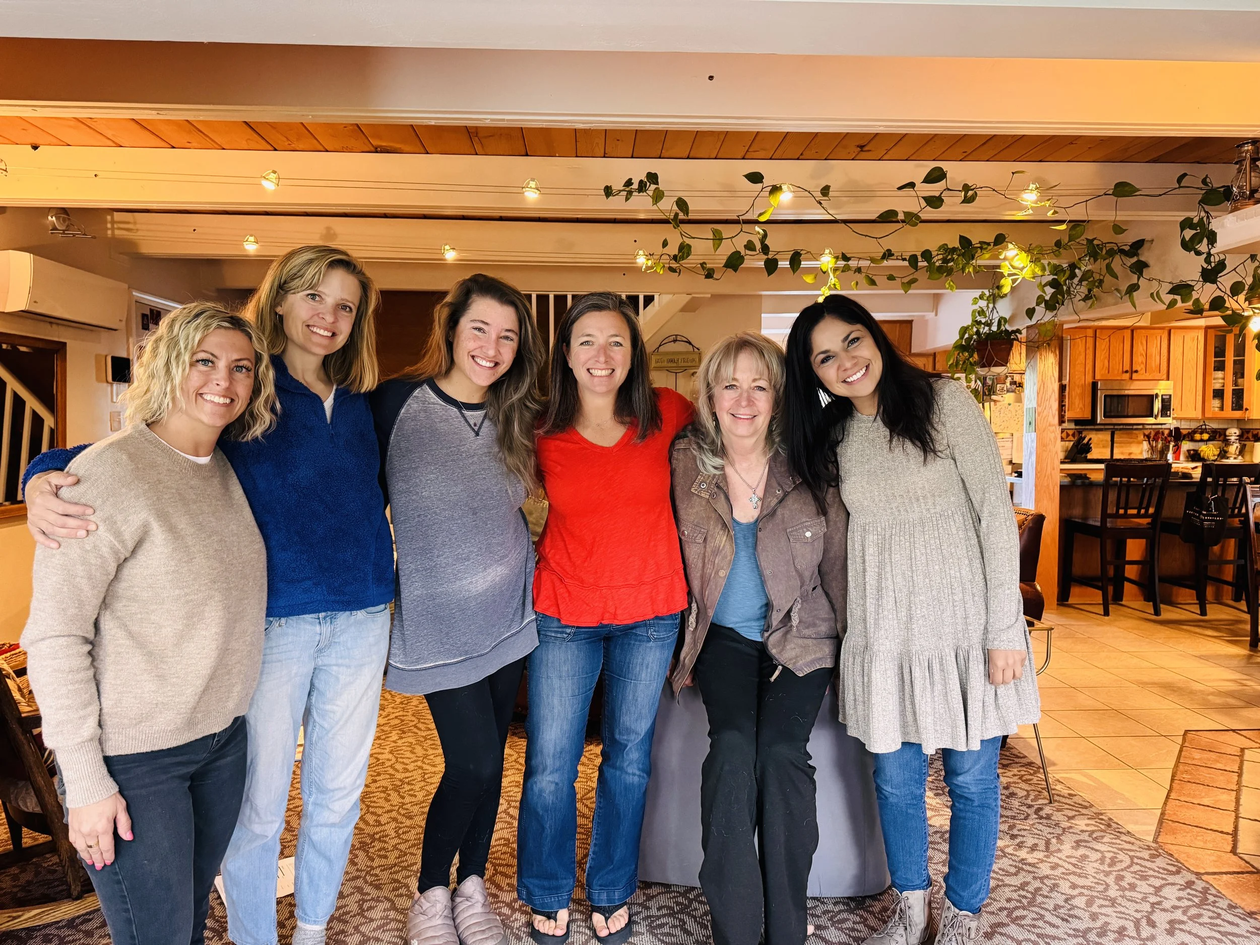 Group of seven women standing closely together inside a cozy home, smiling at the camera.