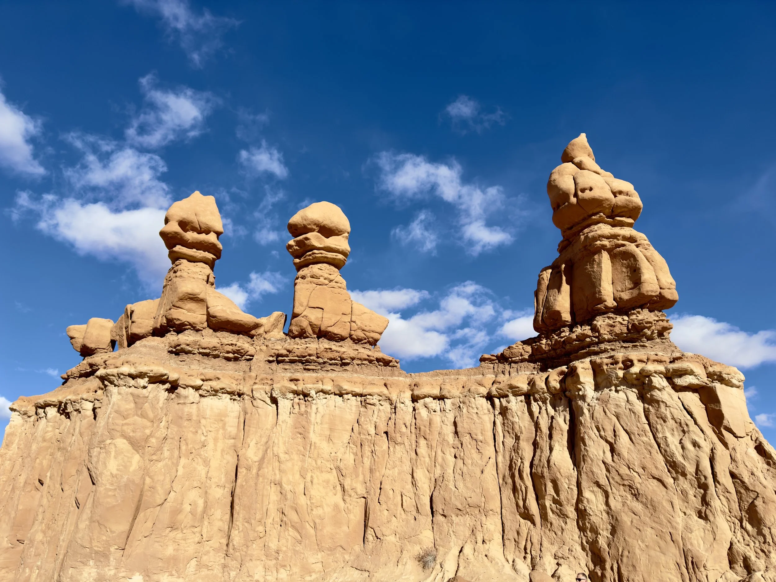 Rock formations with three tall, uniquely shaped spires in a desert landscape against a blue sky with scattered clouds.