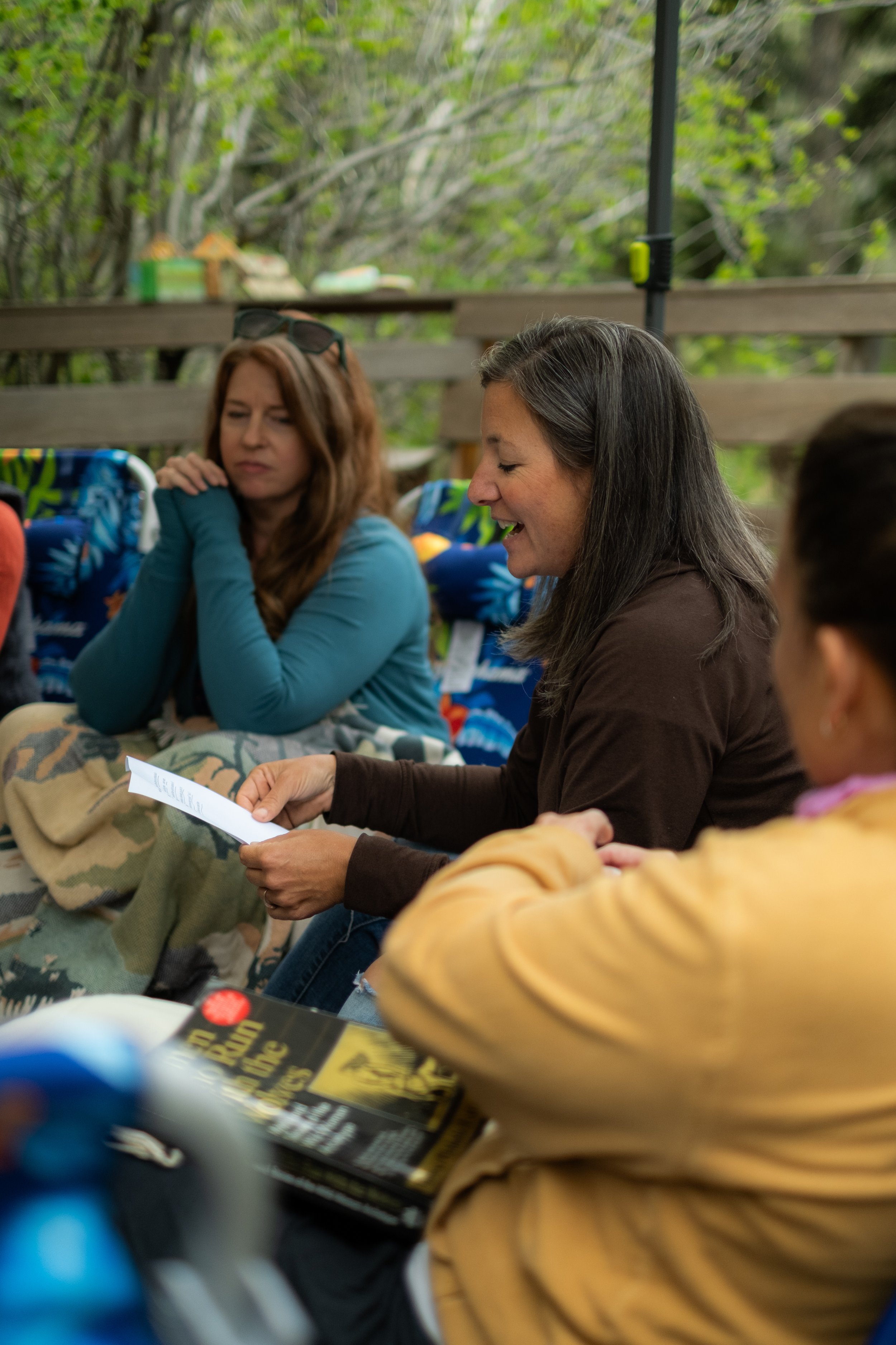 Three women sitting outdoors, engaged in conversation, with books and snacks on the table. One woman with long dark hair is reading a paper, another with red hair and glasses on her head appears to be listening, and a young girl with dark hair is sitting nearby. The background shows green trees and a wooden fence.