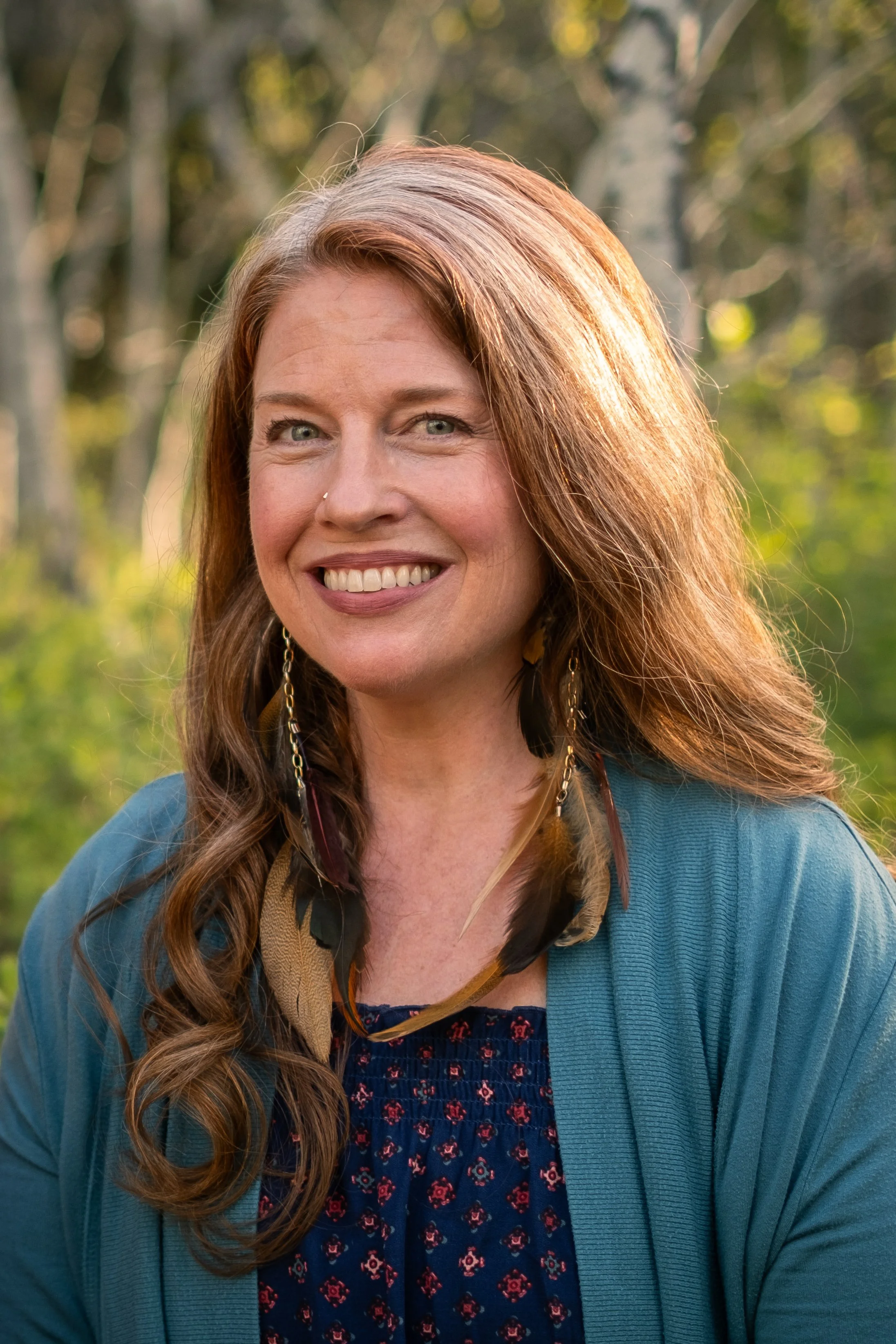 A woman with long, wavy, red hair, smiling, standing outdoors with a blurred background of trees and greenery.