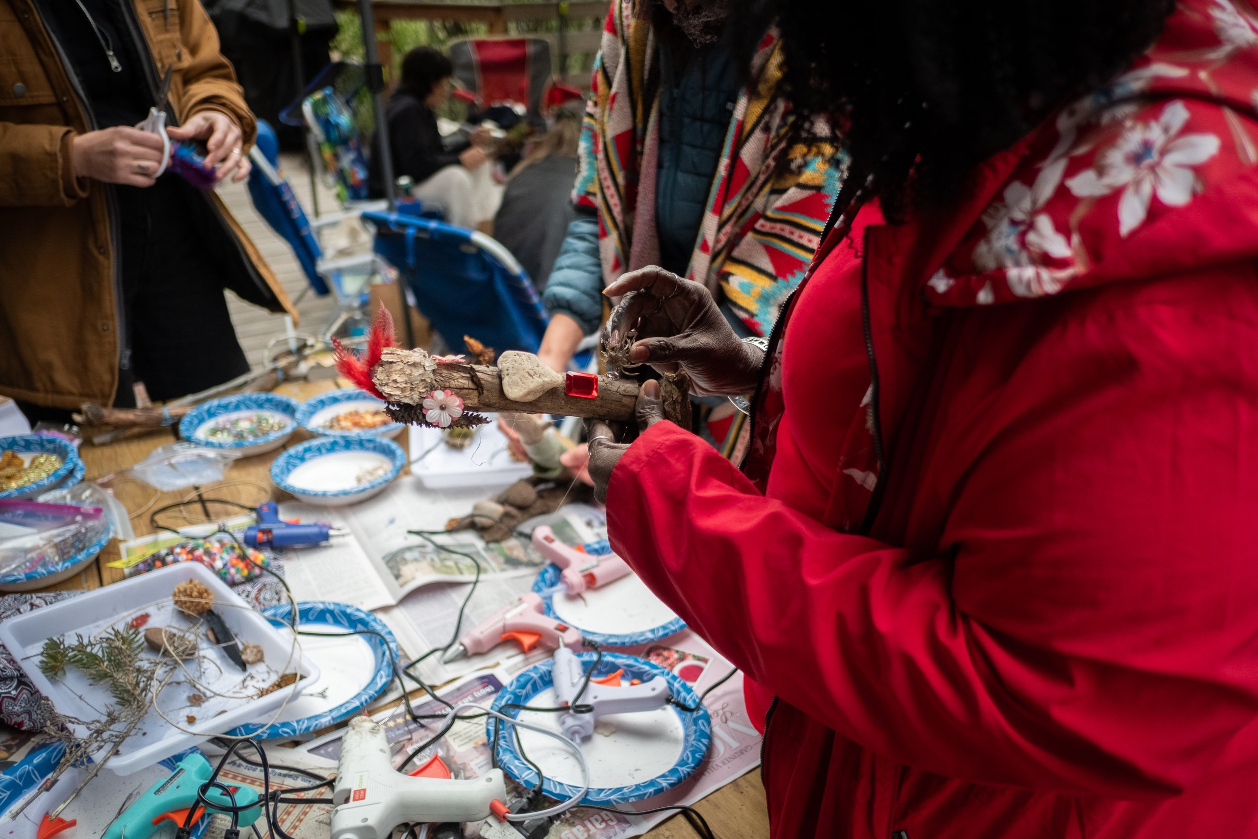 People at a craft table working on handmade art projects with various supplies like glue guns, beads, and natural materials.