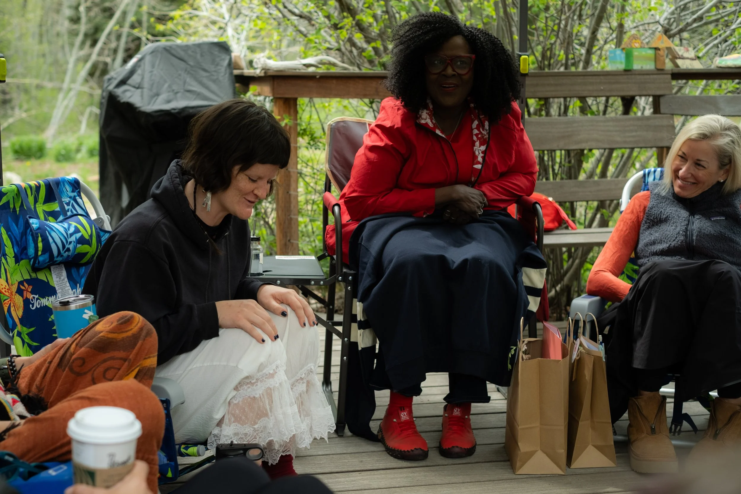 Group of women sitting and standing outdoors, with trees and wooden fencing in the background, during daytime. One woman in the center with curly hair and red glasses is looking forward, others are smiling and engaged with items like papers, bags, and beverages around them.