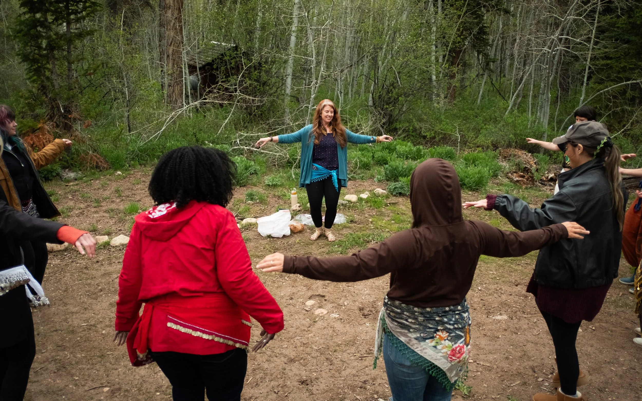 Group of people standing in a circle outdoors in a forest, practicing a yoga or meditation exercise with a woman leading at the center.