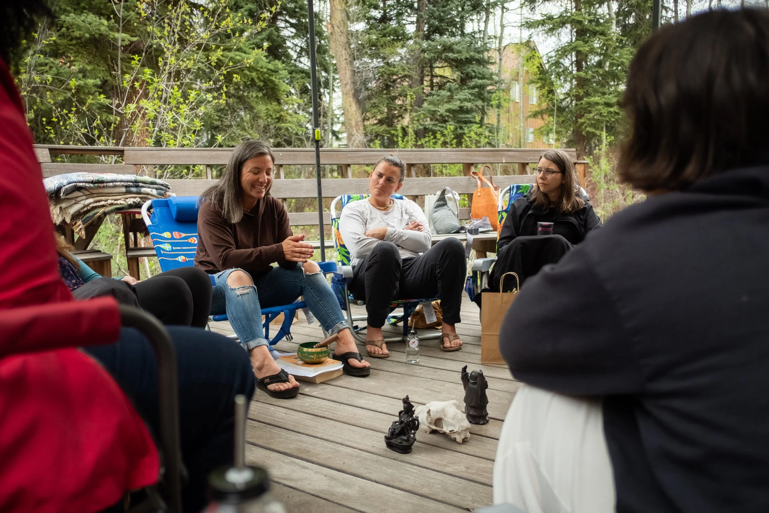 Group of women sitting outdoors on a wooden deck having a discussion, surrounded by trees and nature.