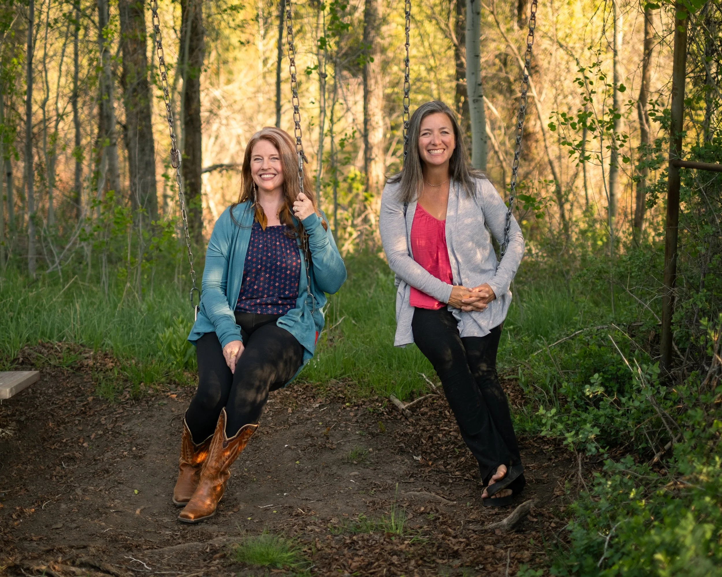 Two women smiling on swings in a wooded outdoor setting, one wearing cowboy boots and a blue jacket, the other in black pants and a gray jacket.